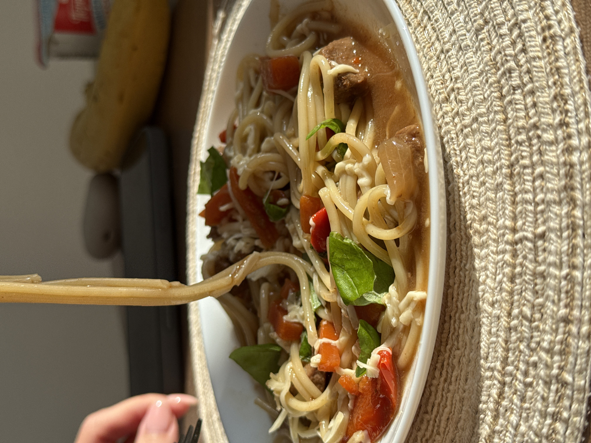 A close-up, rotated, and well-lit image shows a white bowl filled with noodles, red pepper pieces, green leaves (likely basil), and what appears to be meat. The bowl is placed on a woven, tan-colored placemat. A hand holding chopsticks extends into the frame from the lower left, picking up the noodles. In the background to the left are indistinguishable items. The location and city of Perm, Russia, cannot be determined from this image.