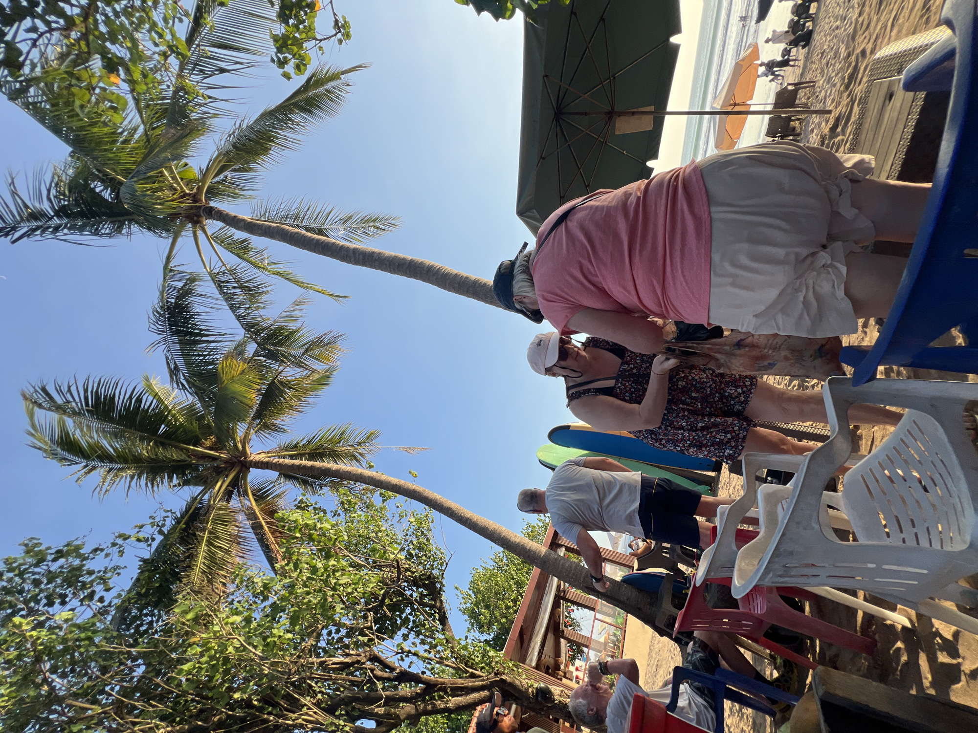 In Kuta, Indonesia, on a sandy beach under palm trees and a clear blue sky, a group of people are gathered near surfboards. The image captures a scene with an ocean in the background, where a few individuals can be seen in the distance. Two women in the foreground are interacting, with one in a pink top and beige shorts, and the other in a floral dress and a light-colored hat. Beside them are several colorful plastic chairs and surfboards leaning against a palm tree. To the right, a large green umbrella provides shade. The overall atmosphere suggests a relaxed, outdoor setting, likely a beachside gathering or waiting area.