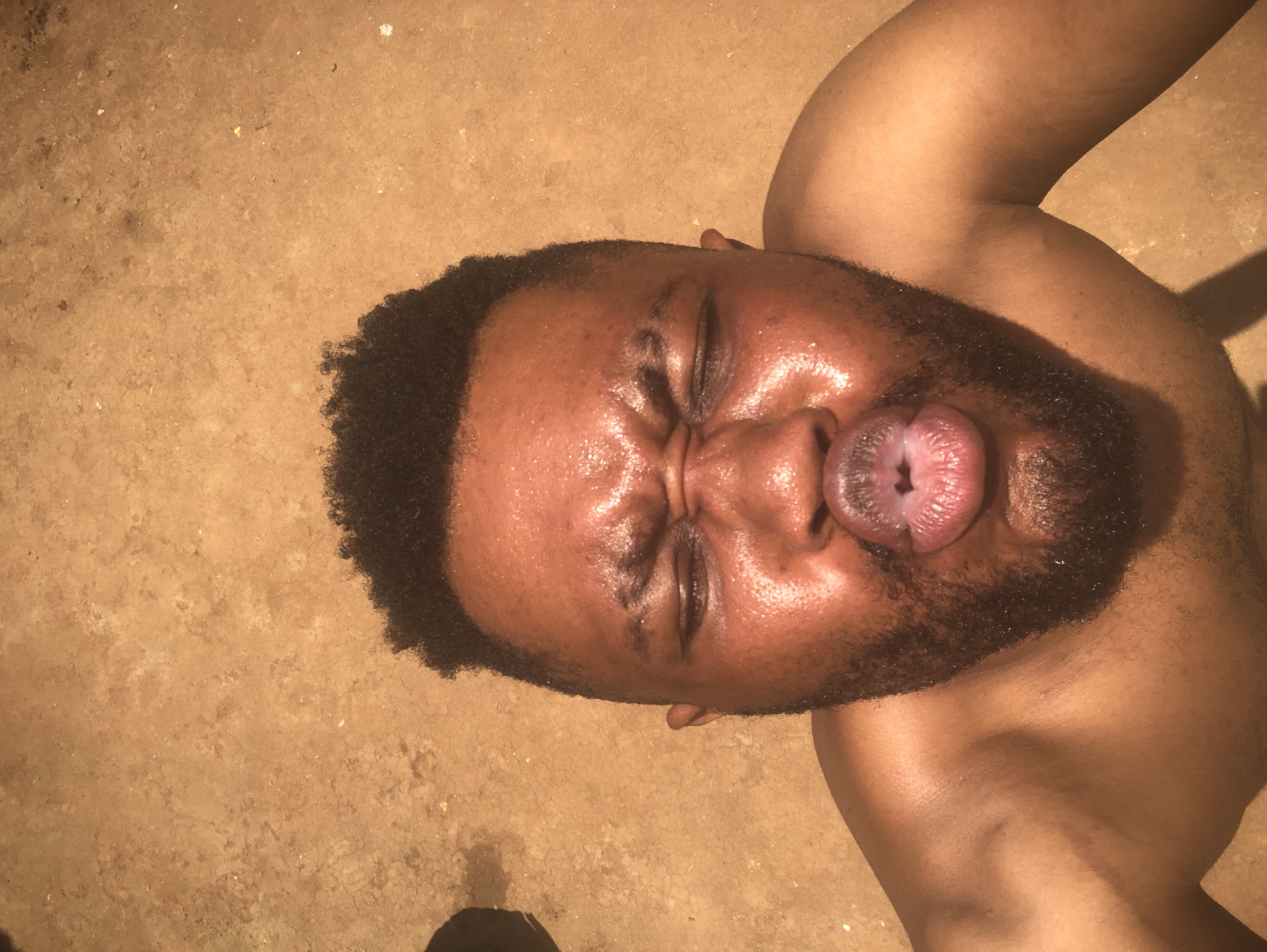 A top-down shot shows a man with dark hair and a beard lying on a cracked, earthen-toned surface. He has his eyes closed and is making a kissing face, extending his lips outward. The man is bare-chested, revealing a portion of his shoulder and upper chest. The location is not specified within the image.