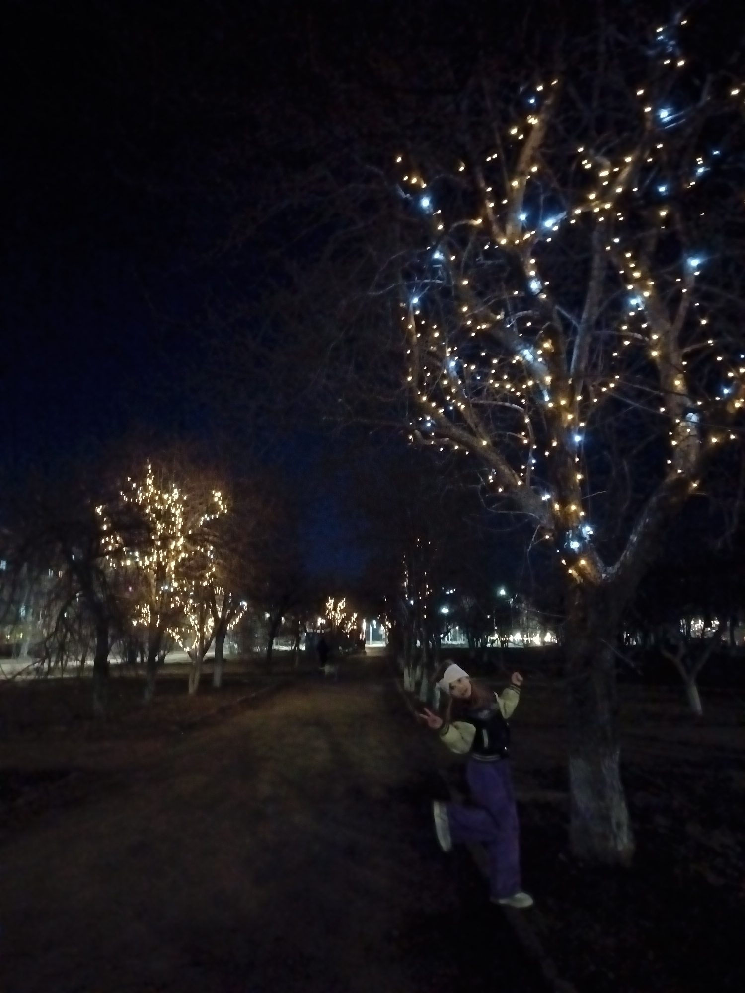 A woman stands at night in a park in Osa, Russia. She is wearing a white hat, a jacket, and purple pants, and is making a peace sign with her left hand. She is standing next to a tree that is decorated with white and gold lights. Behind her are other trees, also decorated with lights, lining a path. The sky is dark. The ground is dirt or grass. The scene suggests it is winter.