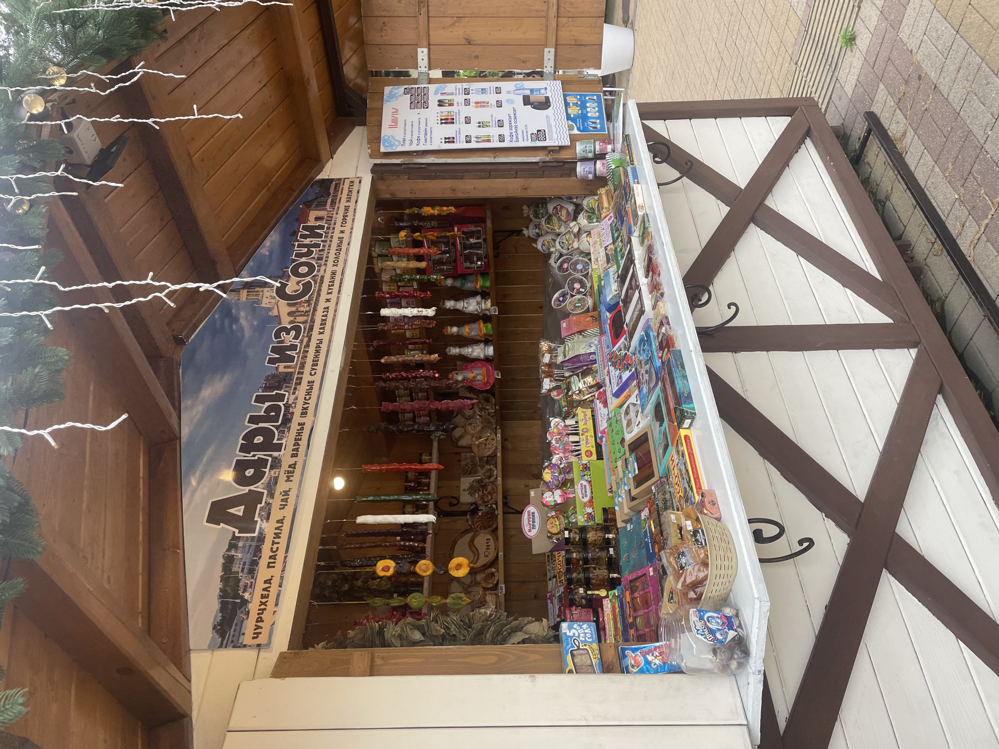 Here is a description of the image:
The image shows a souvenir stand in Sochi, Russia, selling local treats and drinks. The stand is decorated with a sign that reads "Gifts from Sochi" in Russian, and offers "churchkhela, pastila, tea, honey, jam," as well as "cold and hot drinks." Churchkhela, which is a traditional Georgian candle-shaped candy made of nuts and grape juice, is hung on display. Other items for sale include magnets, postcards, and packaged snacks. A price list for various beverages, including coffee and beer, is posted on the stand. Christmas lights are draped around the top of the stand. The stand is partially covered with a wooden roof, and the side has a white wooden lattice design. The background shows a paved sidewalk.