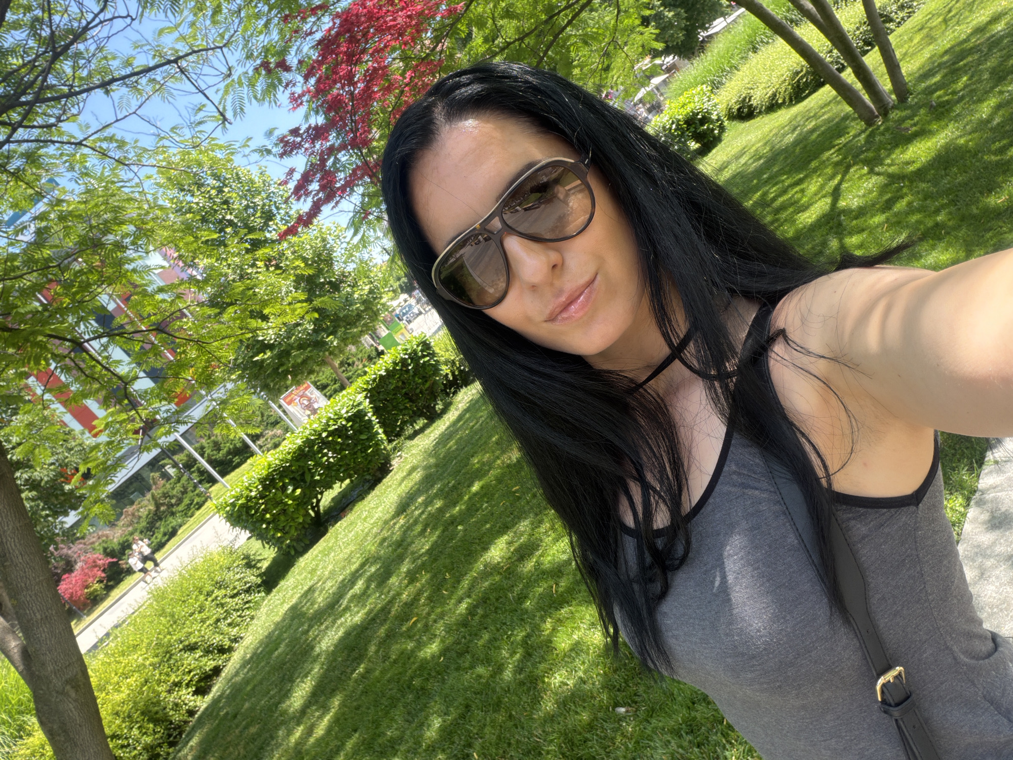 A woman with long, dark hair and sunglasses takes a selfie in a park. She is wearing a gray tank top with black trim and a black strap across her chest. The background shows lush green grass, trees with green and red leaves, and a building with colorful architectural elements, suggesting an urban park setting. The sun is shining brightly, casting shadows on the grass. This scene is located in Pleven, Bulgaria.