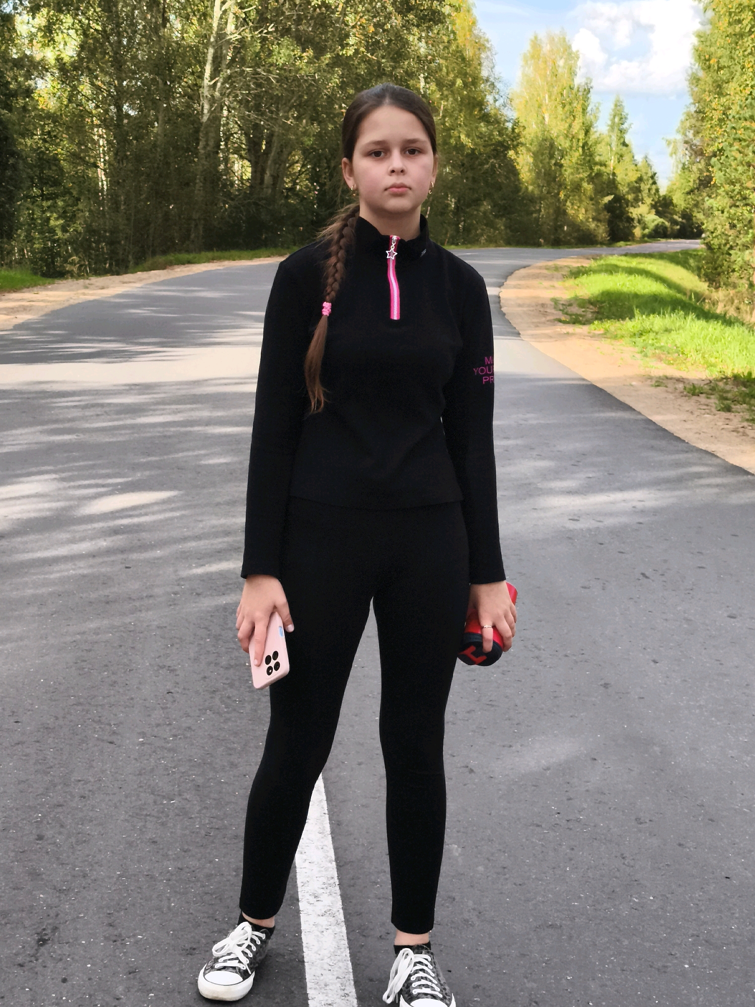 A young girl stands on an asphalt road with trees on both sides. She is wearing a black long-sleeved top with a pink zipper and black leggings. She has a phone in her left hand and a red and black ball in her right hand. Her hair is braided with a pink tie. The road appears to be in a rural area, possibly near Gorki, Belarus, given the presence of trees and the general landscape. The sky is visible above the trees with some clouds.