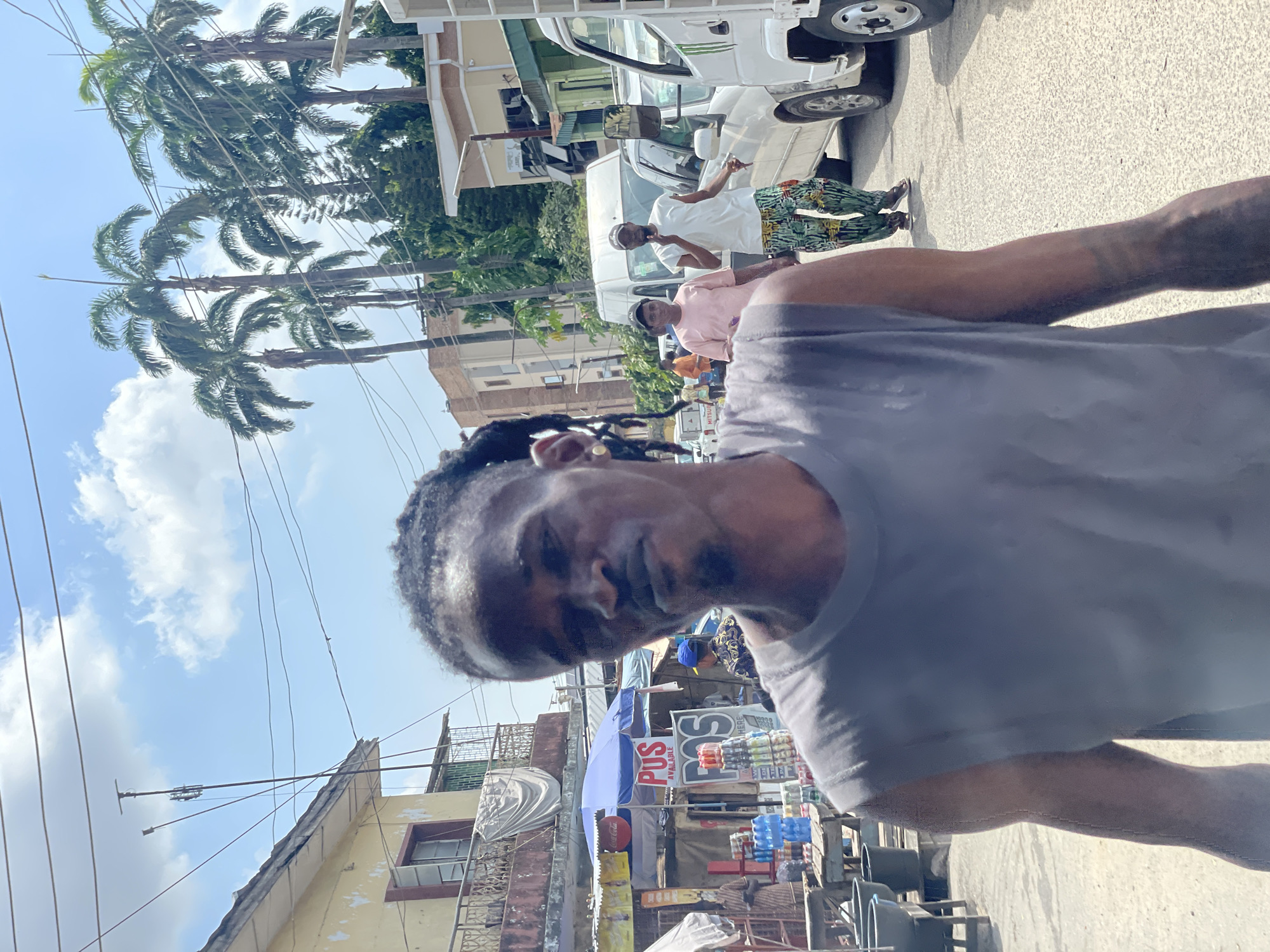 The image depicts a vibrant, sunny street scene in an urban area, consistent with Lagos, Nigeria. The photograph is taken as a selfie by a dark-skinned man with dreadlocks or braids, wearing a grey t-shirt. He looks directly at the camera with a neutral to slightly smiling expression.
In the background, the street is bustling with activity under a bright blue sky dotted with white clouds. Tall palm trees line the left side of the road, their fronds swaying. Numerous overhead wires crisscross the sky above buildings and vehicles.
Several other people are visible on the street. One man wears a white garment with green and black patterned trousers, appearing to be on a phone call. Another person in a light pink top is partially visible. Vehicles, including a white truck or van, are present, suggesting traffic or parking.
To the right, a lively street stall is set up, shaded by a blue umbrella. The stall displays various bottled drinks like water and sodas. A prominent red and white sign reads "POS AVAILABLE HERE" with "PJS" visible above it, indicating the availability of a point-of-sale service. A yellow object with a "Coca-Cola" logo is also seen near the stall. Faintly, "MITSUBISHI" is visible on a vehicle further back.
The surrounding architecture features multi-story buildings, some appearing older with weathered facades, contributing to the character of an established commercial street. The scene conveys the daily energy and commerce of a busy city environment during daylight hours.