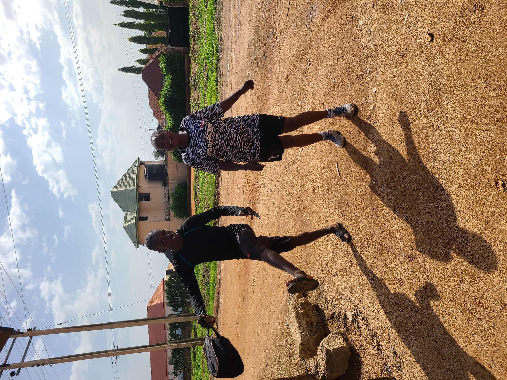 The image captures an outdoor scene in Jos, Nigeria, under a partly cloudy sky. Two individuals, a man and a woman, are the central subjects. The man, on the left, is in mid-stride with his right leg raised, suggesting movement or a playful pose. He is wearing a black long-sleeved shirt, dark shorts, and black sandals. He is also holding a dark bag in his left hand. The woman, on the right, is looking towards the camera with an open-mouthed expression, appearing to be talking or exclaiming. She is wearing a black and white patterned t-shirt with "COOLBET" branding visible, black shorts, white socks, and athletic shoes. The ground is a sandy, reddish-brown dirt path. In the background, several buildings with distinct architectural styles are visible, including a two-story house with a green roof. Greenery, consisting of bushes and tall trees, lines the area behind the subjects. Utility poles and wires are also present in the upper left portion of the image. The lighting suggests it is daytime, casting distinct shadows of the individuals and nearby objects onto the ground. The overall impression is a candid moment captured in a casual, possibly residential, area of Jos.