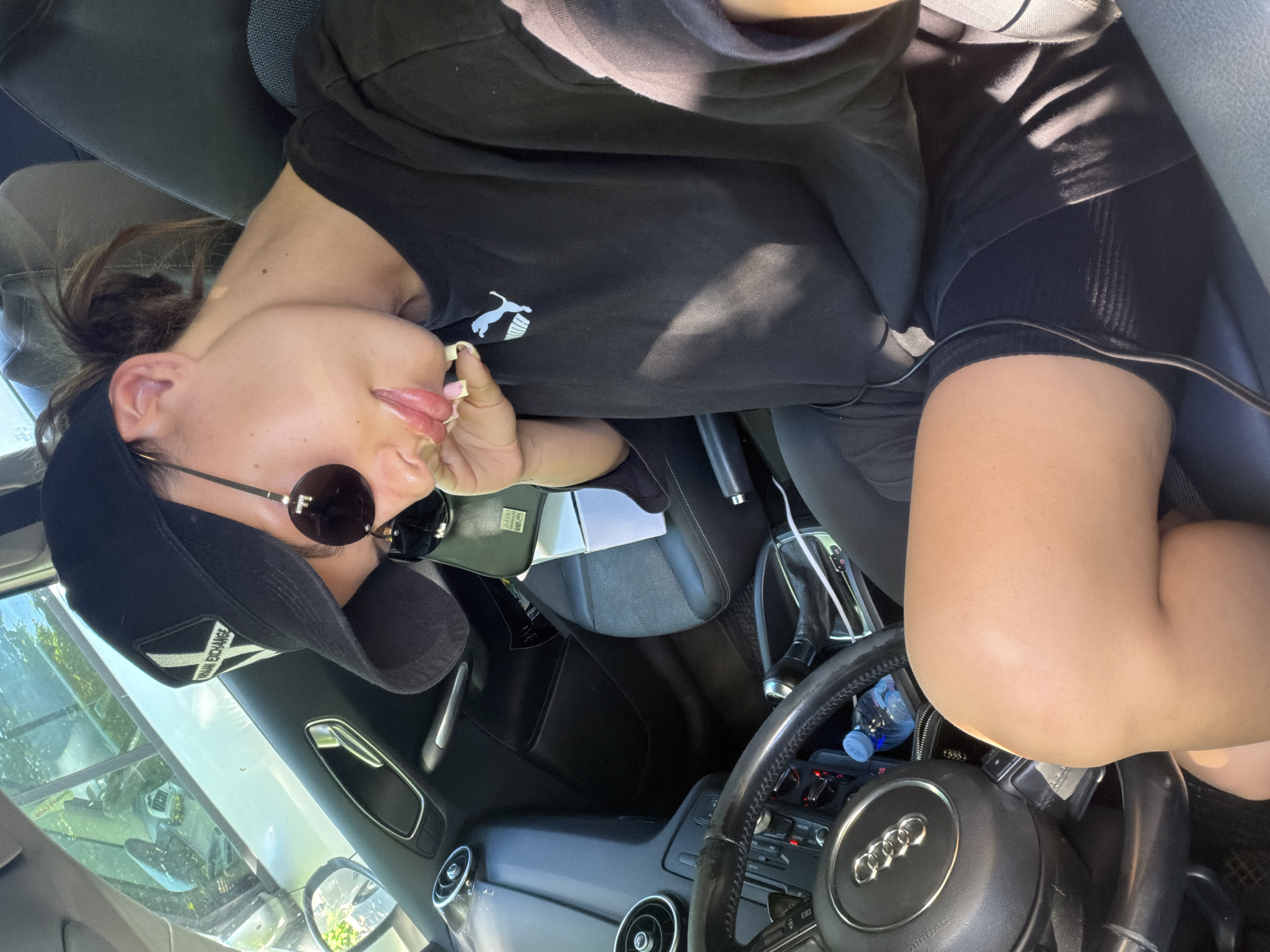 The image shows a young woman sitting in the driver's seat of a car. She is wearing a black baseball cap, sunglasses, and a black Puma sweatshirt over black shorts. Her right hand is resting on her chin, and her lips are slightly parted. The car appears to be parked, and sunlight is streaming through the windows, casting shadows on the interior. The interior of the car shows black leather seats, a steering wheel with an Audi logo, and a gear shift. A water bottle is visible on the center console. The scene is likely set in Burgas, Bulgaria, given the context of the request.