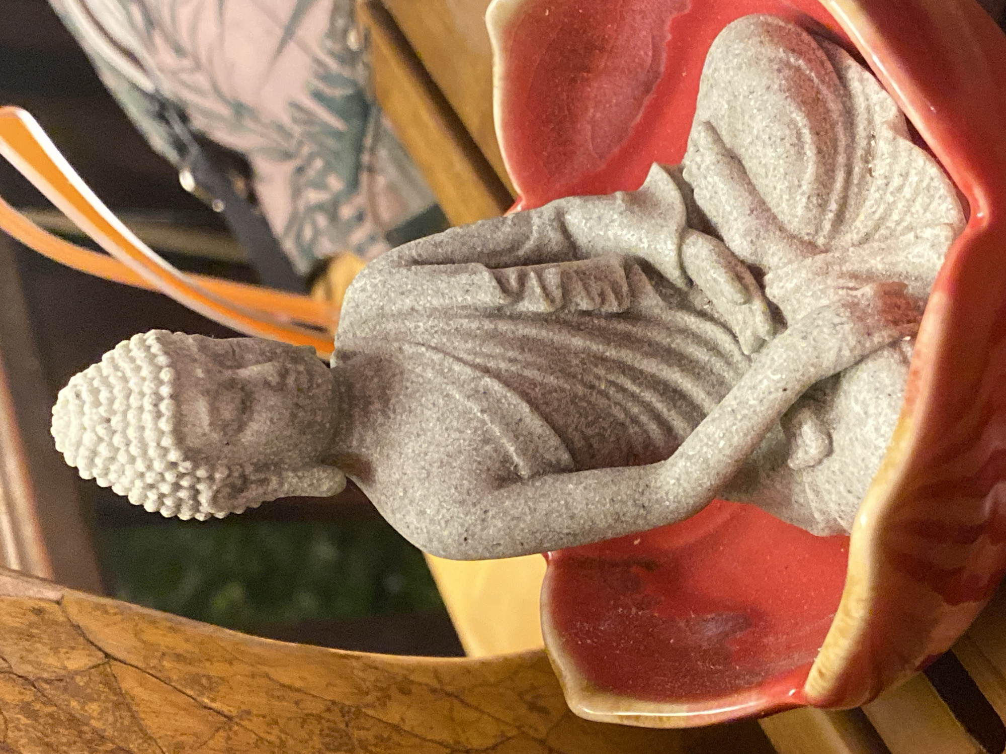 A small, gray, stone statue of the Buddha sits in a bright red, lotus-shaped ceramic bowl. The Buddha is depicted in a seated meditation pose, with its eyes closed and draped in a robe. The statue has a textured, pebble-like surface. The ceramic bowl has a glossy finish and a slightly irregular, organic shape with a pale yellow rim. The bowl is placed on a wooden surface, likely a table, with a slatted texture visible in the background. To the left of the Buddha, a portion of a tall, textured, golden-brown object, possibly made of wood or a natural material, is visible. In the upper right background, a pair of bright orange-rimmed glasses and a person's arm wearing a patterned sleeve are out of focus. The environment appears to be indoors, with soft lighting. The specific location in Baan Mae Nam, Thailand is not discernible from the image itself.