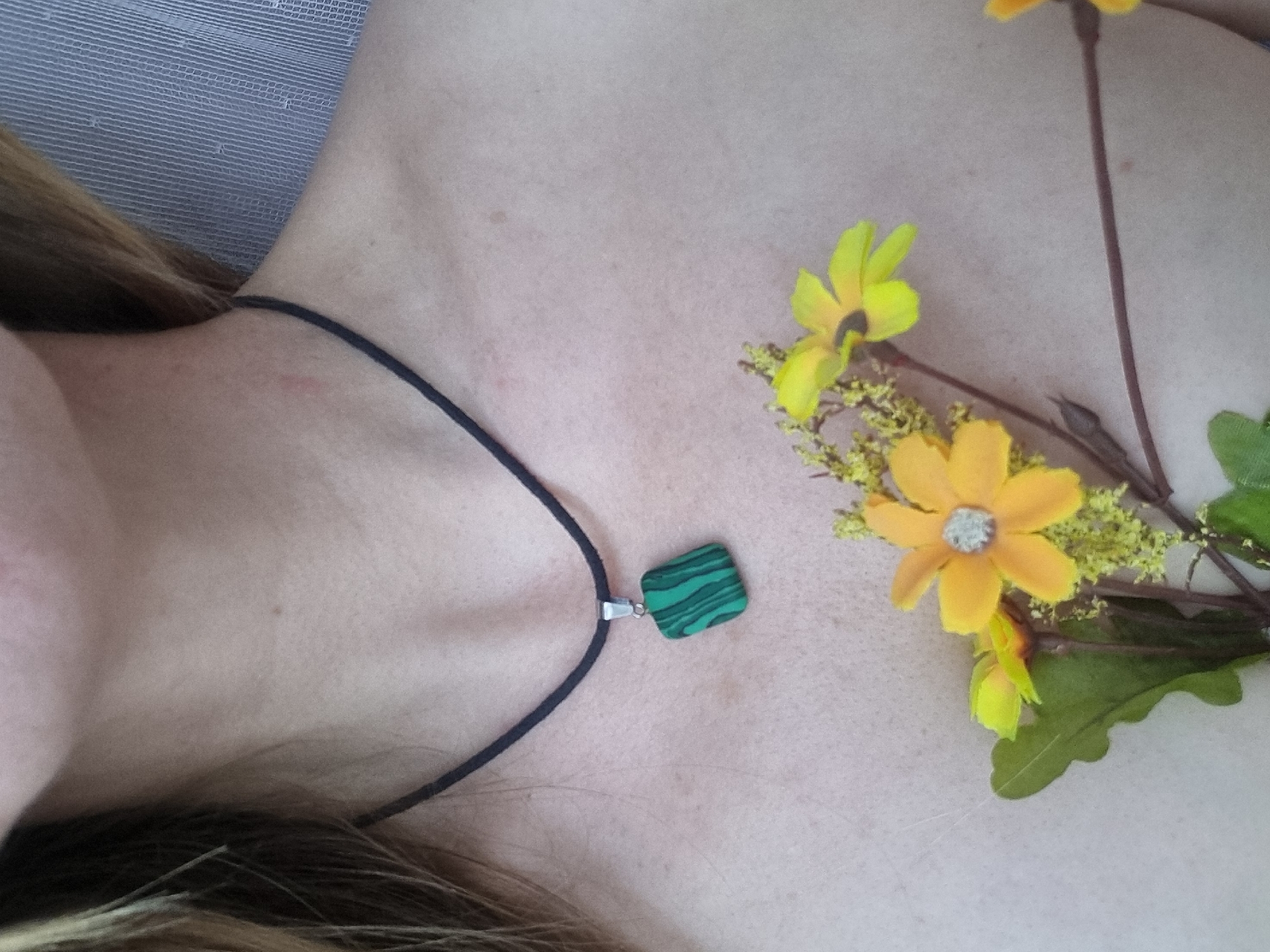 A close-up photograph shows a person's chest and upper neck, adorned with a necklace featuring a green and black striped pendant. Yellow artificial flowers are arranged to the right of the chest. The location and city of Chaikovsky, Russia are not discernible from this image.