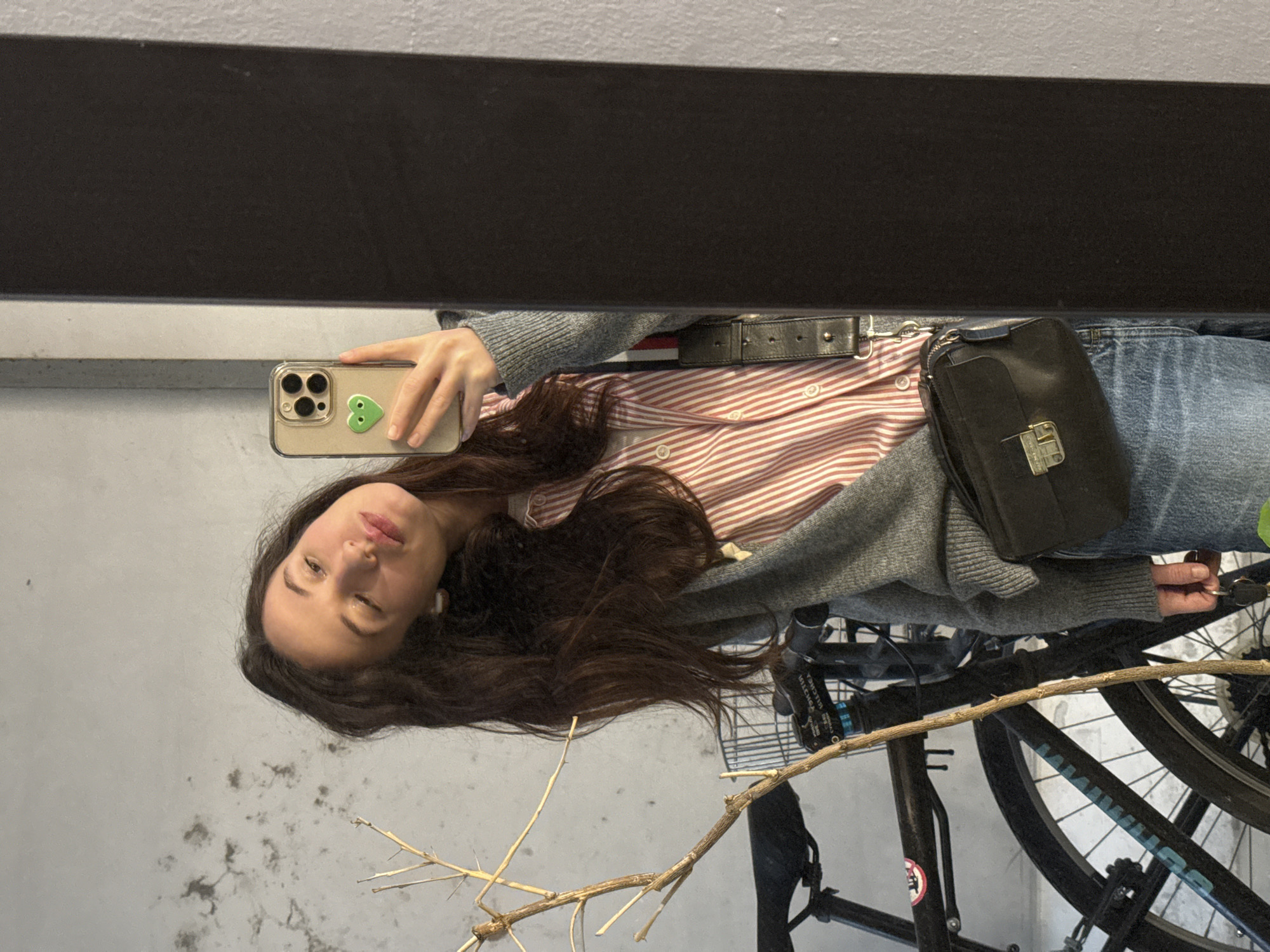 A woman is taking a selfie in front of a mirror. She is holding an iPhone with a green heart sticker on the back. She is wearing a red and white striped shirt, a gray cardigan, and blue jeans. She has long brown hair and is wearing an earbud in her left ear. A black purse is slung across her body, and a black belt is around her waist. In the foreground, there is a bicycle. A dry branch is in the lower center. The background appears to be a gray wall. There is no definitive information to confirm it being in Tel-Aviv, Israel.