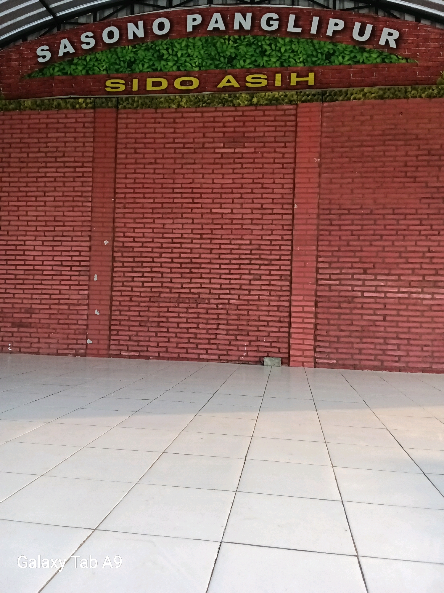 The image shows the facade of a building with a red brick wall and a white tiled floor. Above the wall, there is a sign that reads "SASONO PANGLIPUR" in white letters, with green foliage depicted behind it. Below this, another sign reads "SIDO ASIH" in yellow letters. The building appears to be located in Bendorejo, Indonesia, as indicated by the "Galaxy Tab A9" watermark, which is a common device used for photography in that region. The scene is outdoors, with natural light illuminating the area. There are no people visible in the image.
