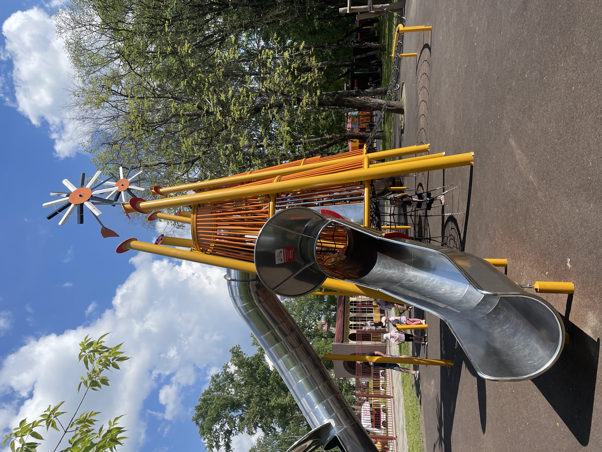 The image shows a children's playground in Vologda, Russia. The playground features a large, modern metal slide, a climbing structure with yellow poles, and a decorative windmill-like element at the top. Several children are visible on the playground, some playing, and others standing. The ground is a dark, rubberized surface. The background includes trees, a fence, and a building with arched windows. The sky is blue with white clouds.