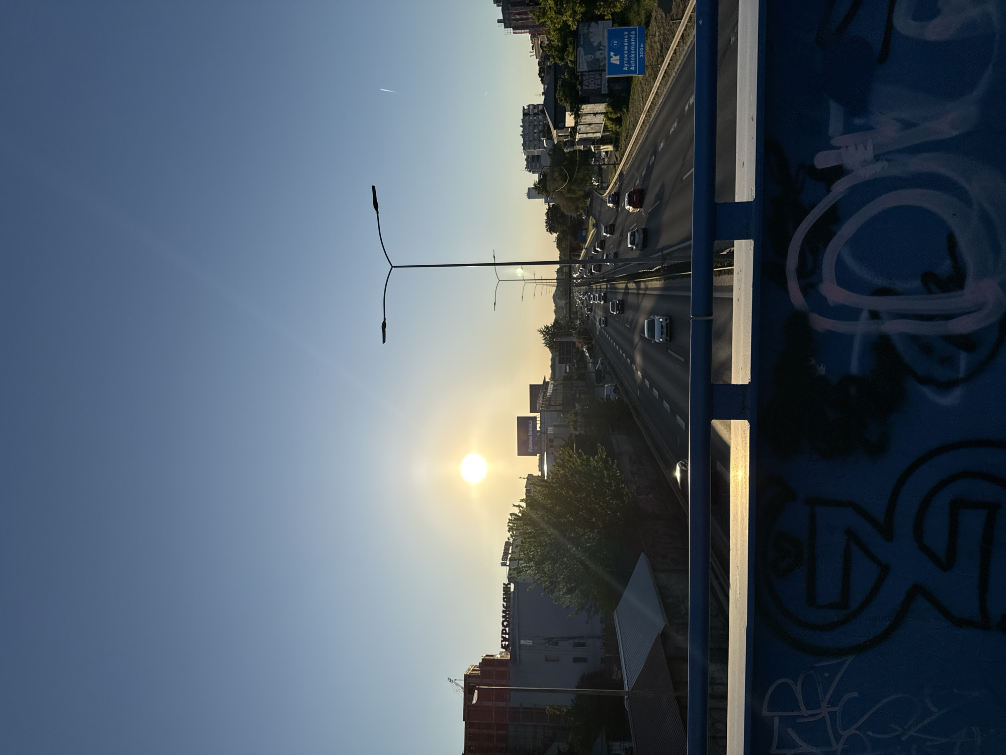 The image shows a view from a bridge overlooking a highway in Vozdovac Urban Municipality, Serbia. The sun is setting, casting a warm glow over the scene. The highway is filled with cars, indicating heavy traffic. On the side of the highway, there are buildings and trees. The bridge itself is visible in the foreground, with blue railings and graffiti. There are light posts along the highway. A sign that reads "Autokomanda" is also visible near the highway.