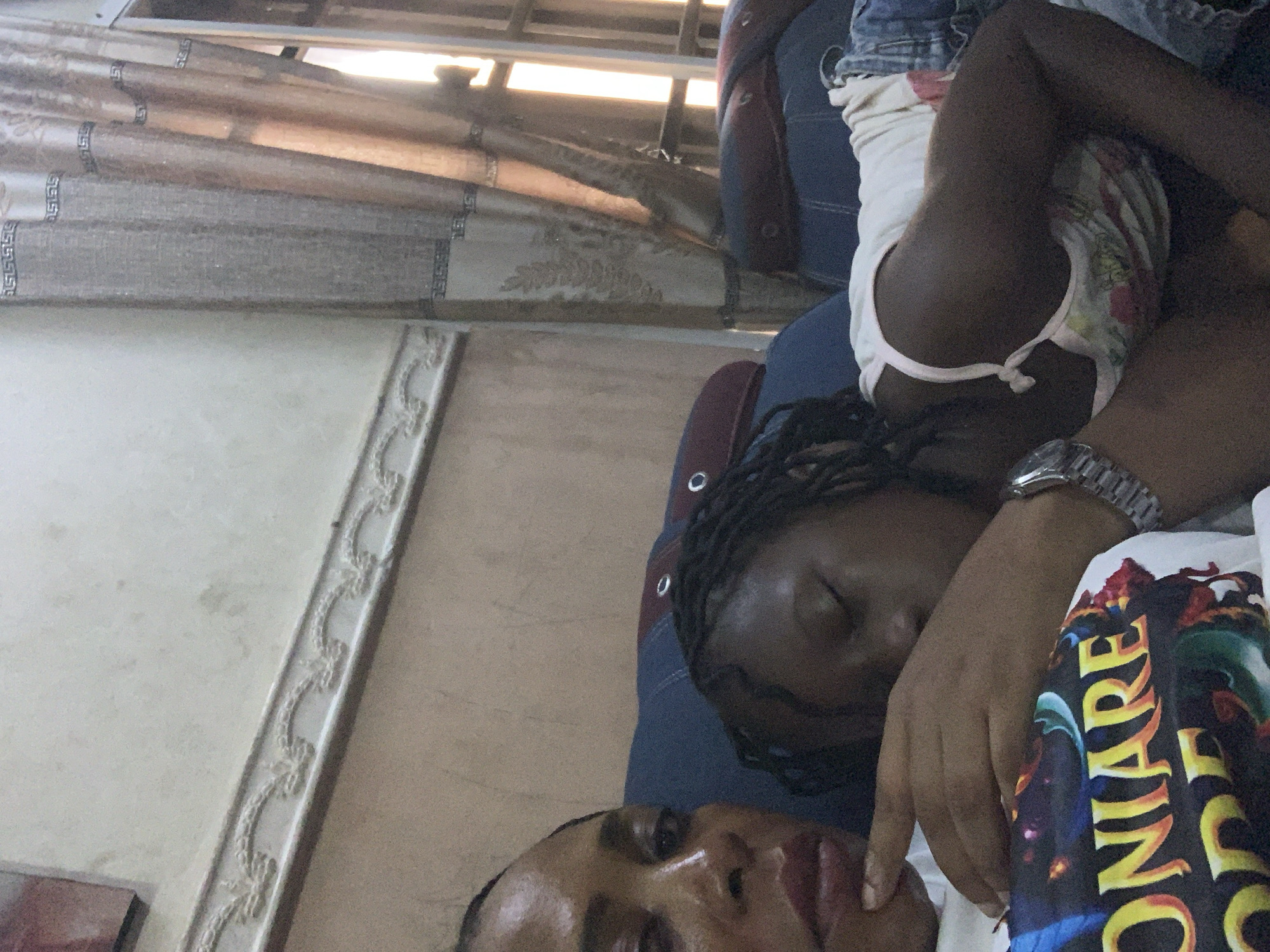 The image shows two individuals, likely a mother and child, resting on a couch in what appears to be a living room in Sabon Tasha, Nigeria. The adult, on the left, is looking towards the camera with a pensive expression. The child, on the right, is asleep, with their head resting on the adult's arm. They are both wearing casual clothing. The background shows a wall with decorative molding and curtains, suggesting an indoor setting. The overall mood is one of quiet repose and intimacy.