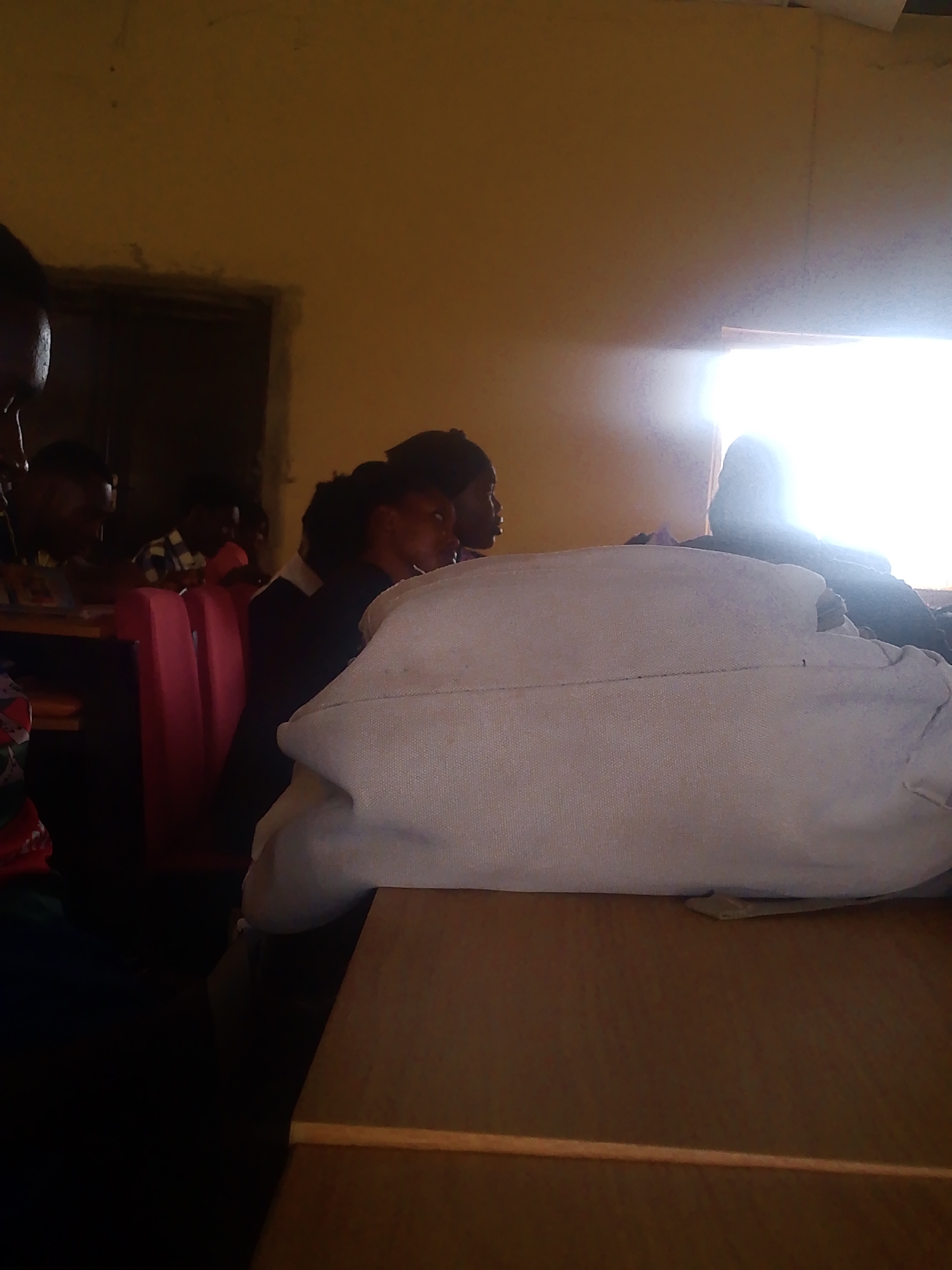 The image depicts an indoor scene, possibly a classroom or a meeting room, located in Gumel, Nigeria. A group of individuals, primarily young adults, are seated at desks, facing towards the right side of the frame. The environment appears to be dimly lit, with a bright light source emanating from a window on the right, creating a strong glare.
In the foreground, a large, light-colored object, possibly a bag or a cushion, is placed on a wooden table. The surface of the table is visible and appears to be made of polished wood.
Behind this object, several people are seated. On the left, the partial profile of a person's face is visible, looking towards the left. Further back, more individuals are seated, with their heads and shoulders visible. One person on the left is wearing a checkered shirt.
The middle ground shows a row of red chairs. Behind these chairs, more people are seated, their faces largely obscured by shadow or other individuals.
On the right side of the image, a bright light source is visible, which could be a window or a light fixture. A person is silhouetted against this light.
The walls of the room are painted a yellowish-brown color. The overall impression is a candid shot of people in a room, possibly engaged in an activity or discussion, with a focus on the immediate foreground and a bright, distracting light source on the right.