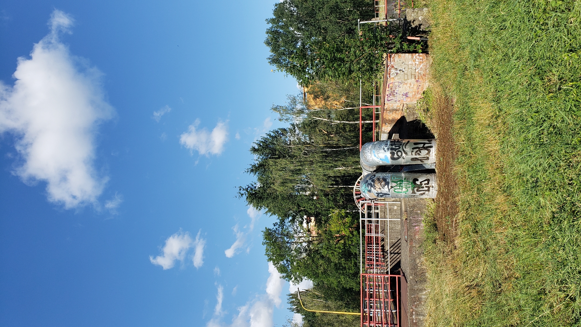 This is a vertical, eye-level, full outdoor shot of a sunny day in Prešov, Slovakia. In the foreground, a grassy area with some dry patches is visible. Behind the grass, a concrete embankment supports two large, graffiti-covered pipes. To the left of the pipes is a red-railed walkway or bridge, with white railings. Beyond the embankment, a dense line of trees forms the background, with some taller trees in the center and shorter ones on either side. A yellow lamppost is visible on the far left. In the distance, behind the trees, a building with an orange facade can be seen. The sky is a clear blue with a few scattered white clouds. The overall scene appears to be an outdoor industrial or public area, possibly near a waterway or water treatment facility.
