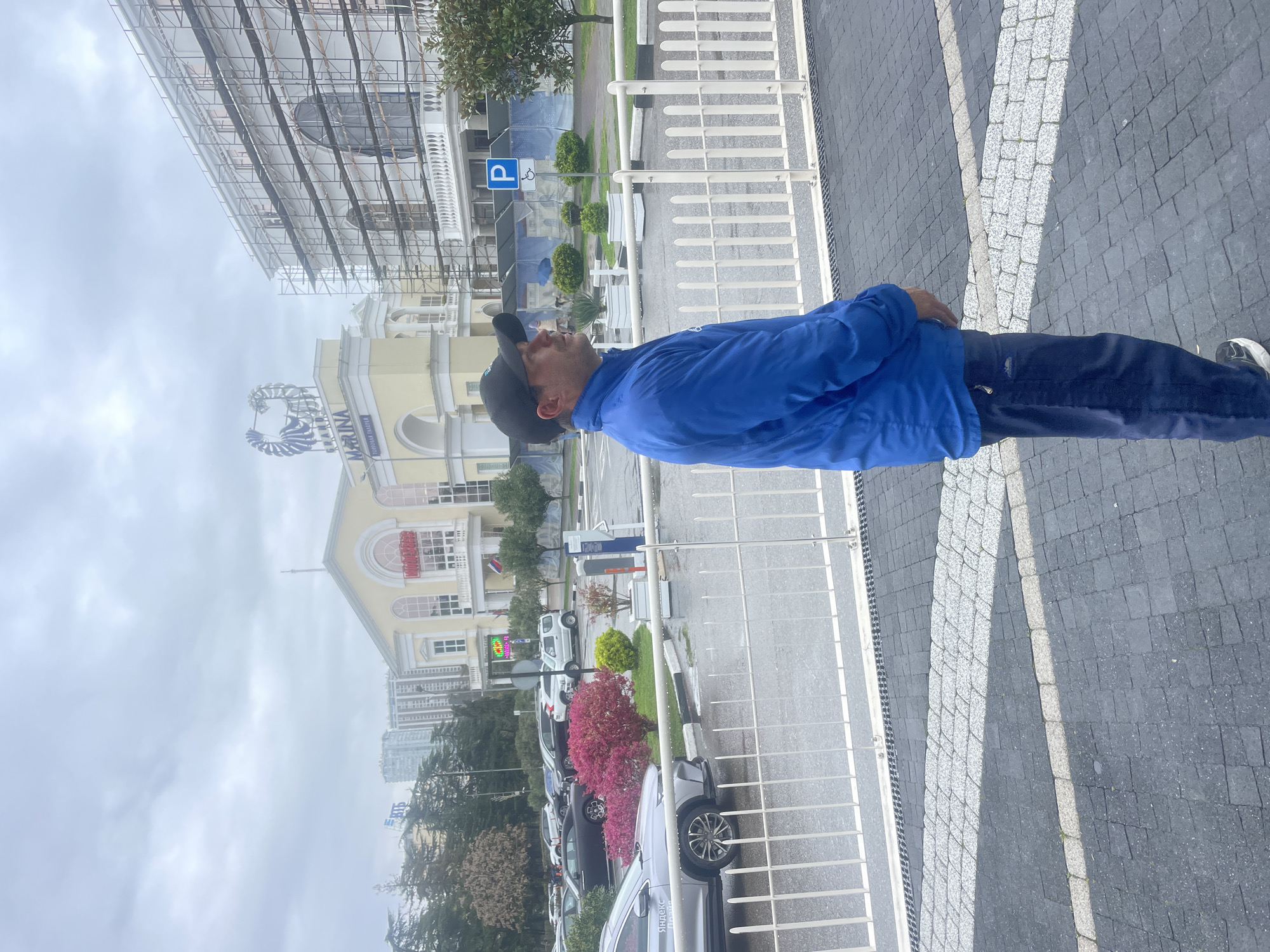 A man in a blue jacket and black baseball cap is standing outdoors on a paved surface next to a metal fence. Behind him is a parking lot with several cars, landscaping with trees and shrubs, and a building with a parking sign. In the background, there is a tall building under construction and the sky is overcast. The location appears to be Sochi, Russia. The building in the background appears to be the Sochi Marina.