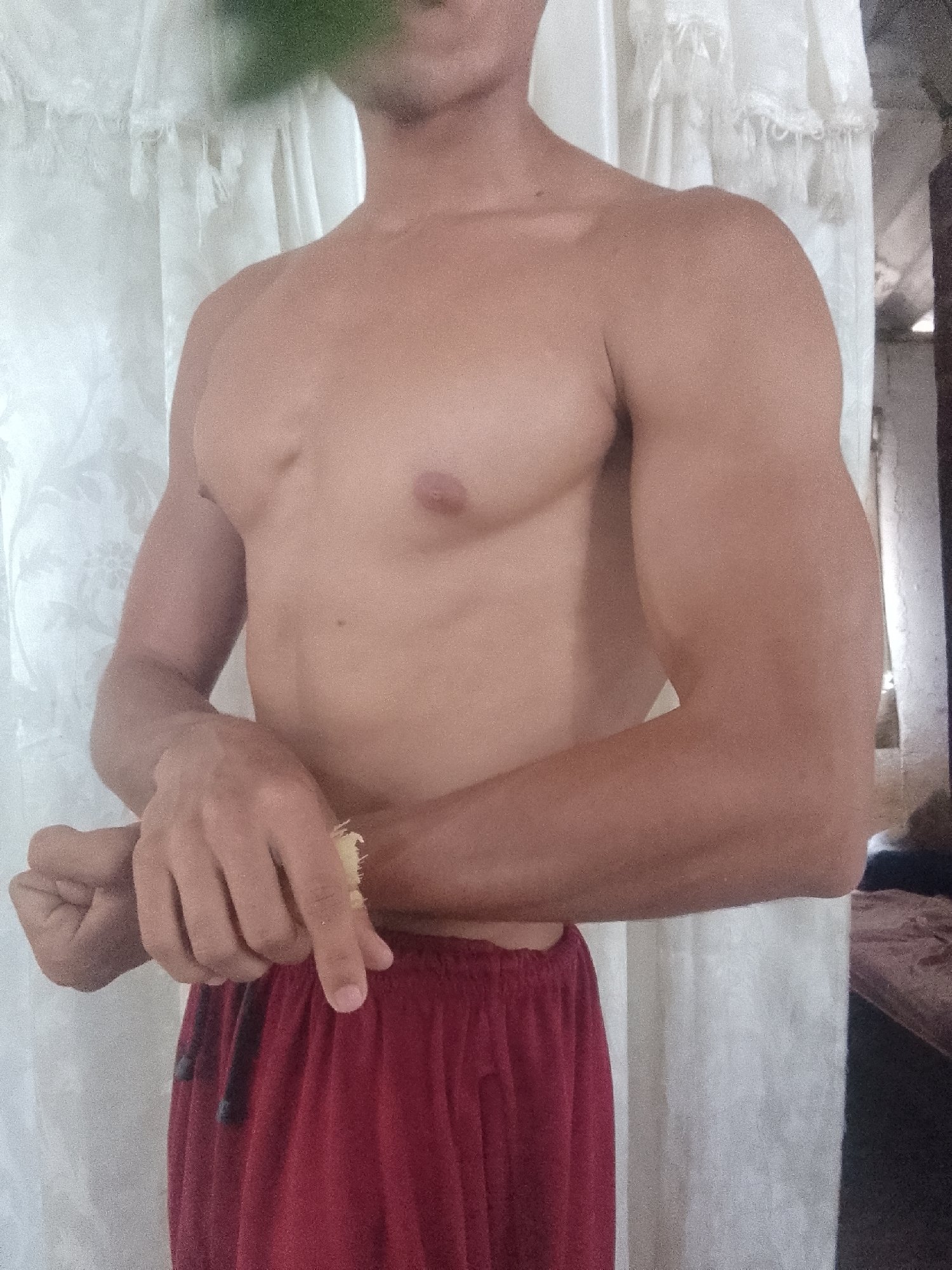A torso of a shirtless man wearing red shorts is positioned in front of a white curtain. The location is Juan Griego, Venezuela, as identified in the prompt. The man's right arm is flexed, and he appears to be examining something small between his fingers. Part of a bed frame and a dark object are visible in the background to the right of the curtain. A blurred green object obscures the top of the frame.