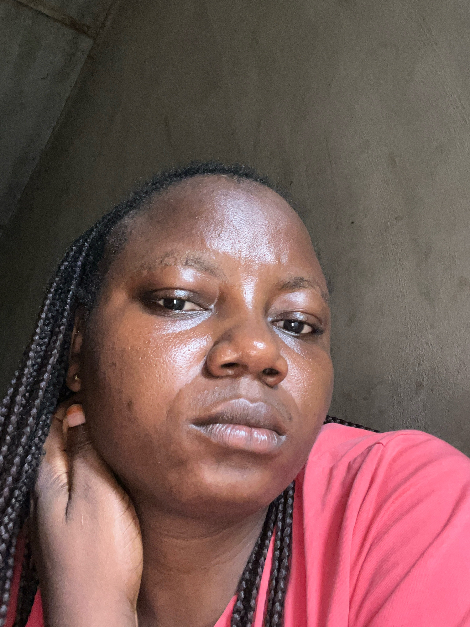 A medium shot captured from a slightly low angle shows a Black woman from the chest up, posed in what appears to be an indoor setting. She is facing the camera directly, with her right hand gently touching her right cheek, and her left hand resting on her neck. Her hair is styled in dark, medium-length braids that extend down her left side, with some braids falling over her shoulder. She is wearing a bright pink t-shirt. The background is a plain, textured wall that is a dark, neutral gray color. The lighting appears to be coming from the left side of the frame, illuminating her face and creating highlights on her skin. The overall mood of the image is somber, with the woman's gaze directed intently at the camera. The image is set in Ikotun, Nigeria, and the scene depicts a personal moment captured within a domestic or private space.