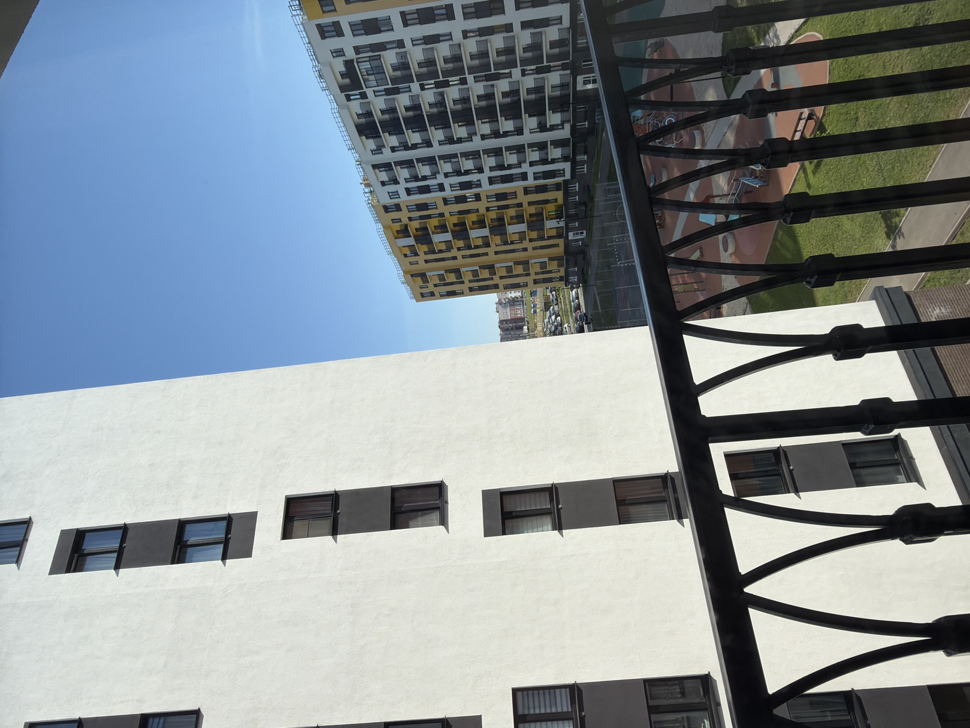 The image, taken from a high vantage point possibly within an apartment building, shows a view overlooking an urban environment. In the foreground is a wrought-iron balcony railing. The midground features two apartment buildings: a white building with horizontally oriented, rectangular windows set in dark grey recesses and a taller, multi-story building with alternating white and yellow sections and numerous windows, possibly indicative of individual apartments. Beyond these buildings, the cityscape stretches into the distance, including what appears to be a parking area with multiple cars and some greenery, indicative of a park or recreational space. The sky is a clear, light blue. Based on the description provided, the location is identified as Pervomaysky, Russia, although the image itself offers no explicit proof of this location. There are no visible people or interactions within the frame.