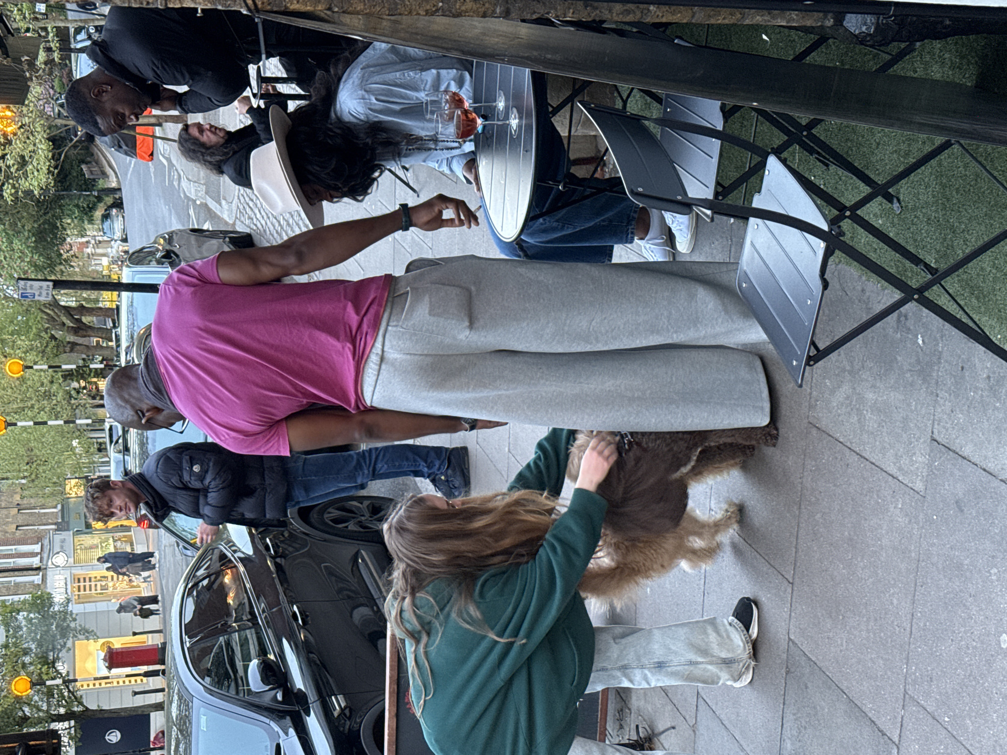 The image captures an outdoor scene in London, United Kingdom. Several individuals are present. A man in a pink shirt and grey sweatpants is standing near a black car. A woman in a green hooded sweatshirt is petting a medium-sized brown dog. Nearby, people are seated at a restaurant with outdoor seating. The table has drinks. A man in a black shirt leans towards the table. The environment includes typical London street elements such as a red mailbox, street signs, and buildings.