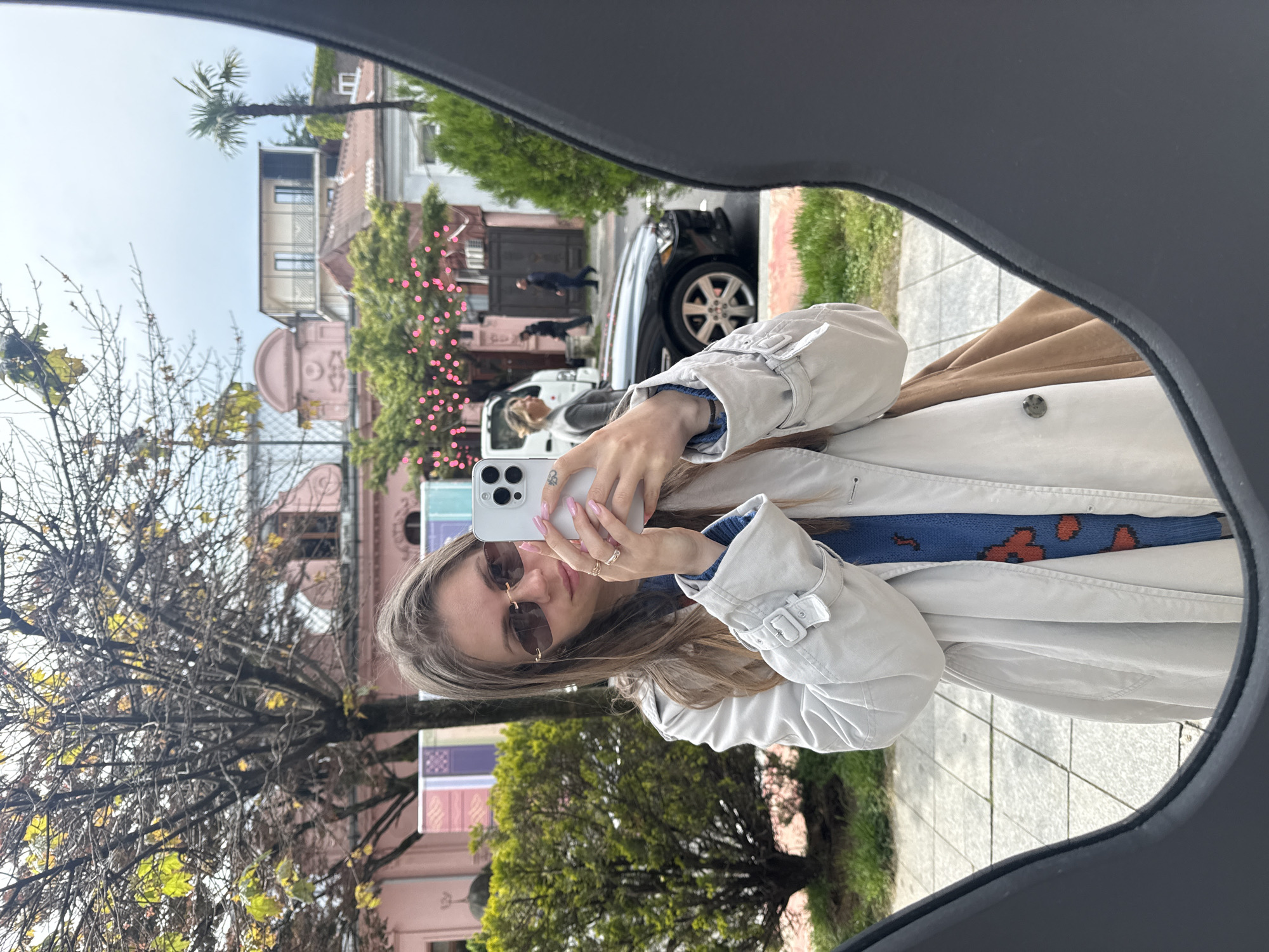 A woman is taking a selfie using a white iPhone, with the reflection visible in a car's rearview mirror. She wears sunglasses and a light-colored coat over a blue sweater. The background shows a street scene in Batumi, Georgia, featuring a pink building adorned with pink floral decorations, trees, a car, pedestrians, and typical urban street elements.