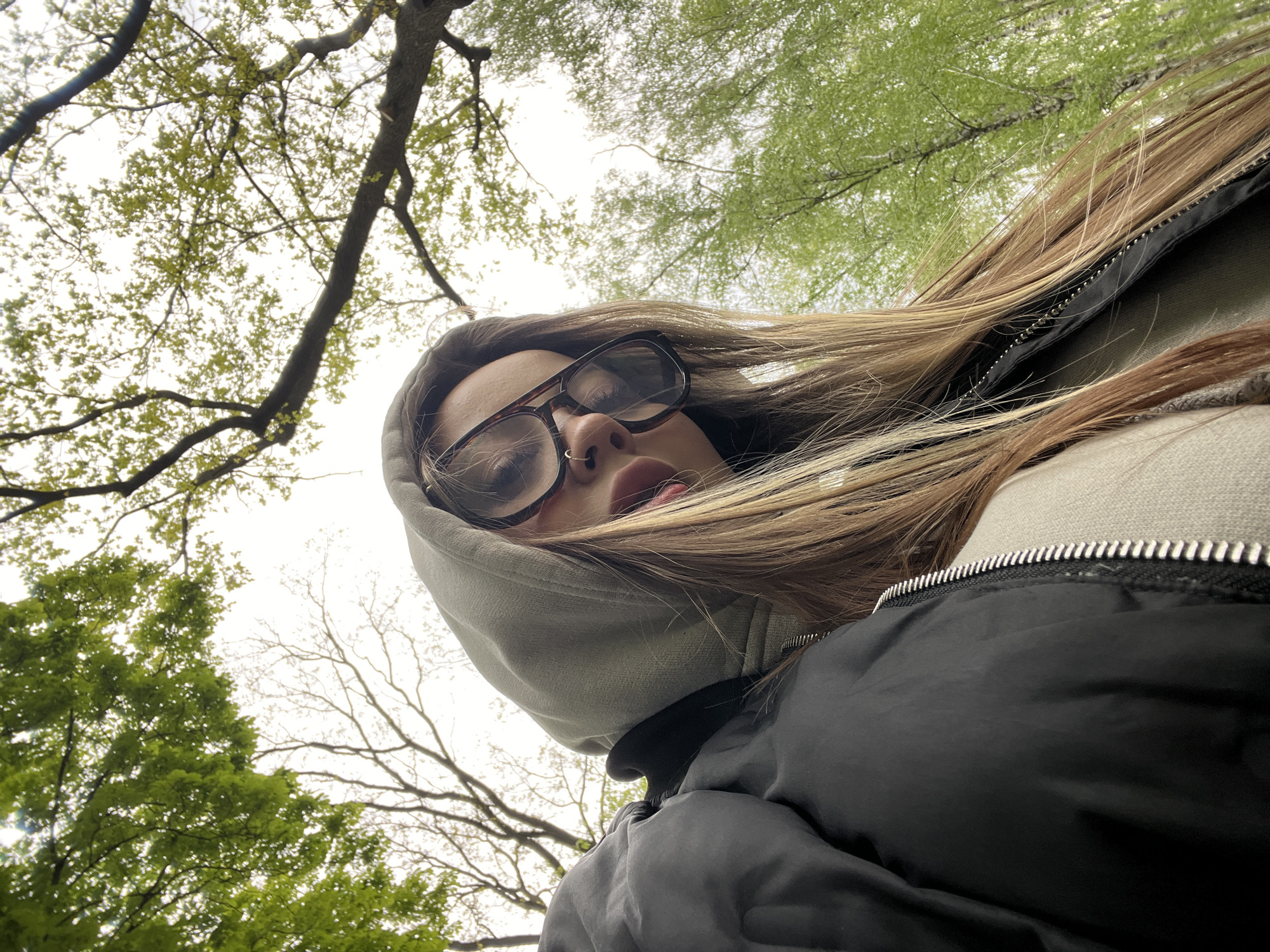 A young woman wearing glasses, a gray hoodie, and a black jacket is looking up at the camera. Her hair is a mix of blonde and brown. The background shows a view of trees and sky, suggesting she is outdoors. I cannot identify that she is specifically in Krasnodar, Russia from just this photograph.