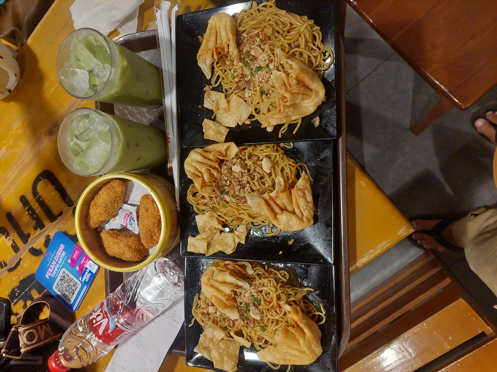 This overhead shot captures a meal at what appears to be a casual eatery. The table is a bright yellow, with a wooden table leg visible on the right. On the table, there are three black square plates, each holding a serving of noodles topped with fried wontons and what looks like minced meat and crispy fried onions. Beside the plates, a bamboo steamer basket contains four breaded, fried items, possibly spring rolls or chicken nuggets, with small packets of sauce. Two tall glasses filled with a green, icy beverage, likely matcha or avocado juice, stand to the left. A bottle of Vit water and a receipt are also present.
A blue QR code sign with Indonesian text, "PESAN DISINI TANPA ANTRI" (Order Here Without Queueing), suggests a self-service ordering system. The overall scene implies a relaxed mealtime, perhaps lunch or dinner. The lighting is bright, suggesting it could be daytime, and there are no obvious cues about the weather. The presence of the QR code and the type of food points towards a modern, casual dining establishment in Indonesia, potentially Pematang Siantar, as indicated by the water bottle brand. A person's feet are visible in the lower right corner, indicating someone is seated at the table. The atmosphere seems laid-back and inviting, focused on enjoying the food and drinks.
