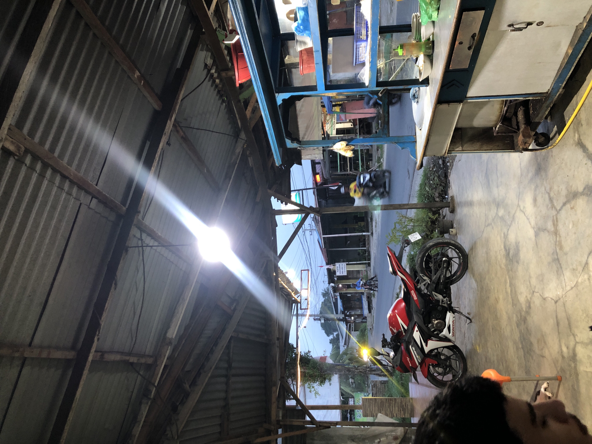 In Sei Mencirim, Indonesia, a motorbike is parked under a covered outdoor seating area, which has a corrugated metal roof supported by wooden beams. The area appears to be a food stall or restaurant. A red and white Honda CBR motorcycle is positioned in the foreground. Beyond the covered area, a street is visible with a blue food cart on the right side of the image. Several people are seen on motorcycles and bicycles on the street, and buildings line the opposite side of the road, including what appears to be a pharmacy or medical store with signage in Indonesian. The lighting suggests it is either early morning or late evening.