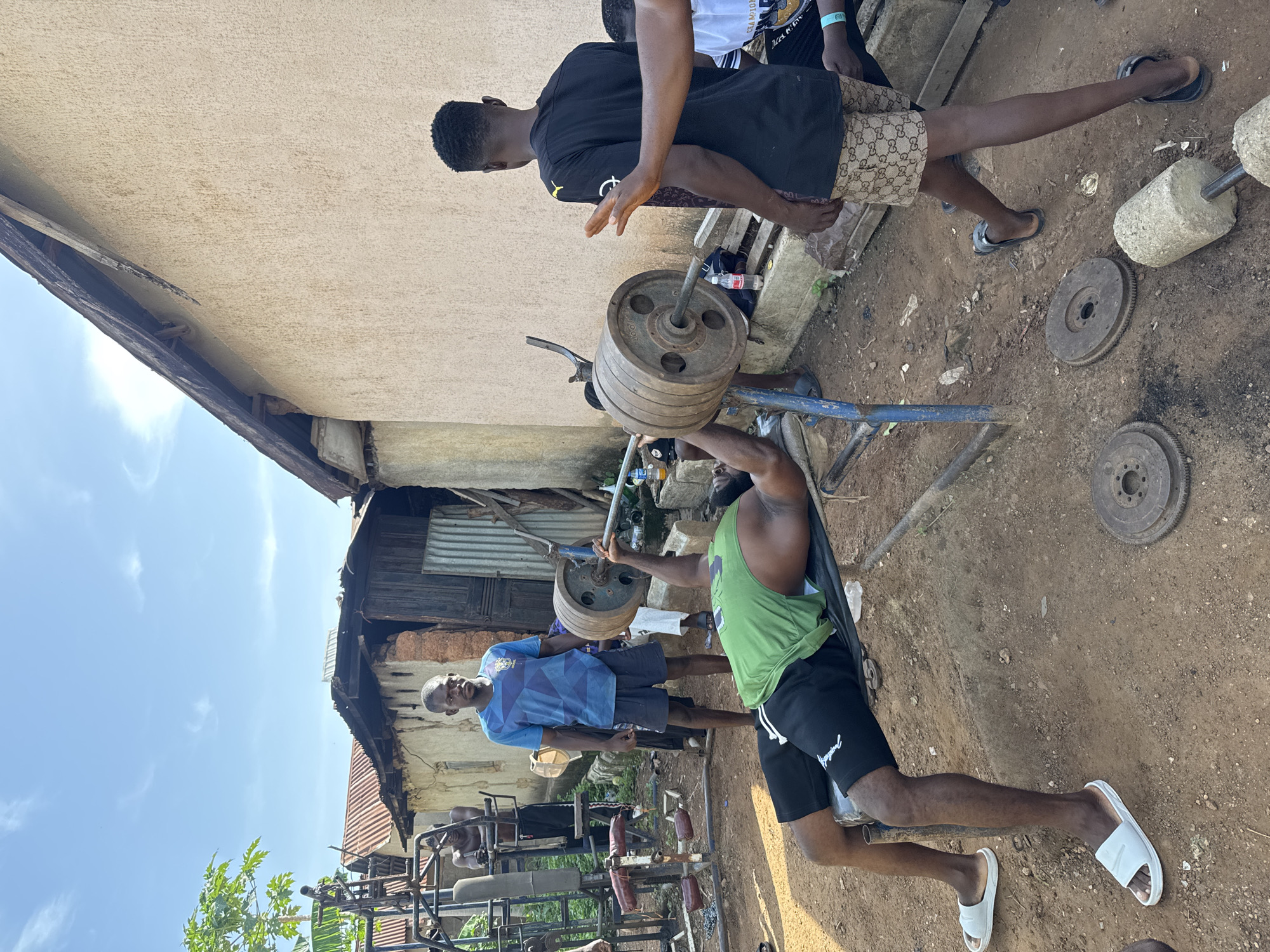 This image, rotated 90 degrees clockwise, captures an outdoor weightlifting session under a bright, clear sky. In the foreground, a dark-skinned man in a green tank top and black shorts with "IMPERIAL" text is performing a bench press on a makeshift blue metal bench. A heavy barbell, laden with numerous iron plates, is at the top of his lift.
Two other dark-skinned men are present, acting as spotters or observers. To the left of the lifter's head, a man in a black t-shirt and patterned shorts (resembling Gucci print) stands ready to assist with the barbell. Further back and to the right, another man in a blue patterned t-shirt and dark shorts watches the lifter intently. A person wearing a white shirt with "CHAMPION" partially visible is in the background near the first spotter.
The setting is an informal, outdoor gym with a dirt ground. Various pieces of gym equipment, including additional weight plates (some appearing to be industrial discs) and concrete weights, are scattered around. In the background, there are simple structures made of plaster and corrugated iron sheets, suggesting a rural or semi-urban environment. Green foliage from a tree is visible on the far left. The bright sunlight indicates it is daytime.