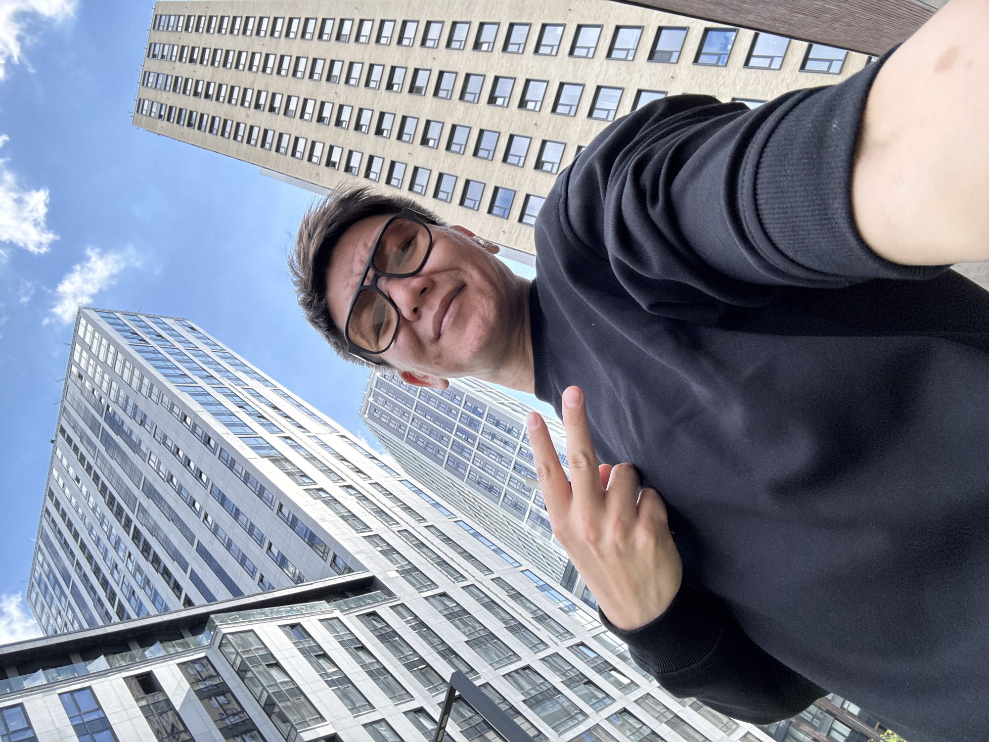 The media file shows a low-angle selfie taken outdoors in an urban environment, likely Moscow, Russia, as indicated by the architecture. The primary subject is a person from the chest up, wearing a black sweatshirt and glasses, looking at the camera with a slight smile. The person is making a peace sign with their right hand, with the index and middle fingers extended.
The background is dominated by tall, modern buildings with repetitive window patterns, typical of city skylines. One building is a light beige color with many dark rectangular windows, while another, to the left and closer to the viewer, is a lighter, more contemporary design with a glass facade. The sky above is blue with scattered white clouds, suggesting a clear day, possibly late morning or early afternoon given the lighting.
The perspective is from below, looking upwards, which emphasizes the height of the buildings and the sky. The scene conveys a casual and optimistic mood, with the subject appearing relaxed and happy. There is no visible text in the image. The focus is on the individual in the foreground against the backdrop of the urban landscape.