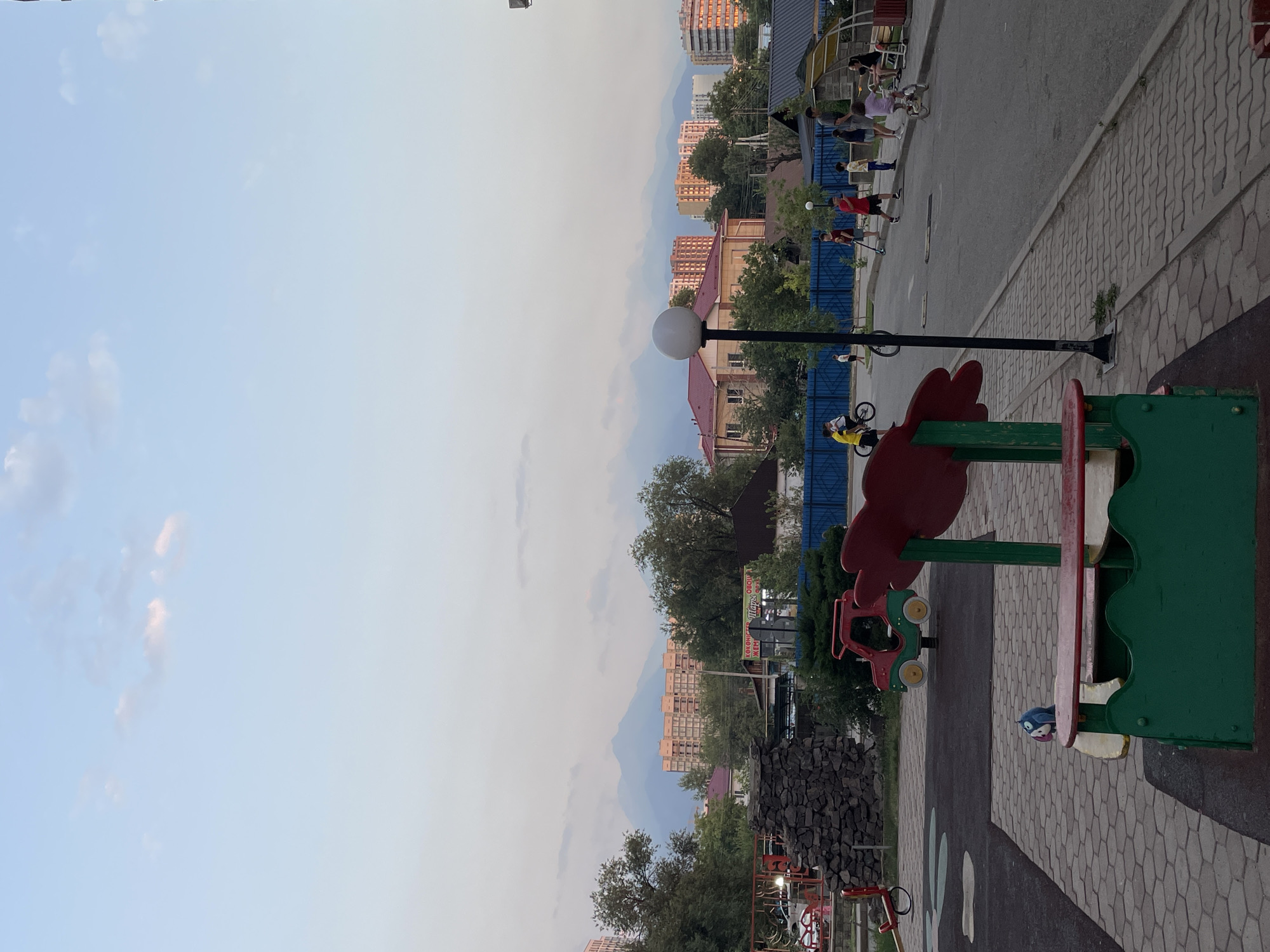 This image captures a residential courtyard in Almaty, Kazakhstan, at dusk. In the foreground, a colorful children's playground structure, predominantly green and red, is visible. To the left, a red and green toy car and a red seesaw are also part of the playground equipment. The ground is paved with interlocking grey stones.
In the mid-ground, several children are engaged in various activities on a paved road. One child rides a bicycle, another is on a scooter, and others are walking. A lamppost stands prominently on the right side of the road. The background reveals a cluster of residential buildings, including multi-story apartment complexes, interspersed with trees. In the far distance, the majestic, mist-shrouded mountains that are characteristic of Almaty's geography can be seen under a pale blue sky with a few wispy clouds. The overall scene suggests a peaceful evening in a neighborhood setting.