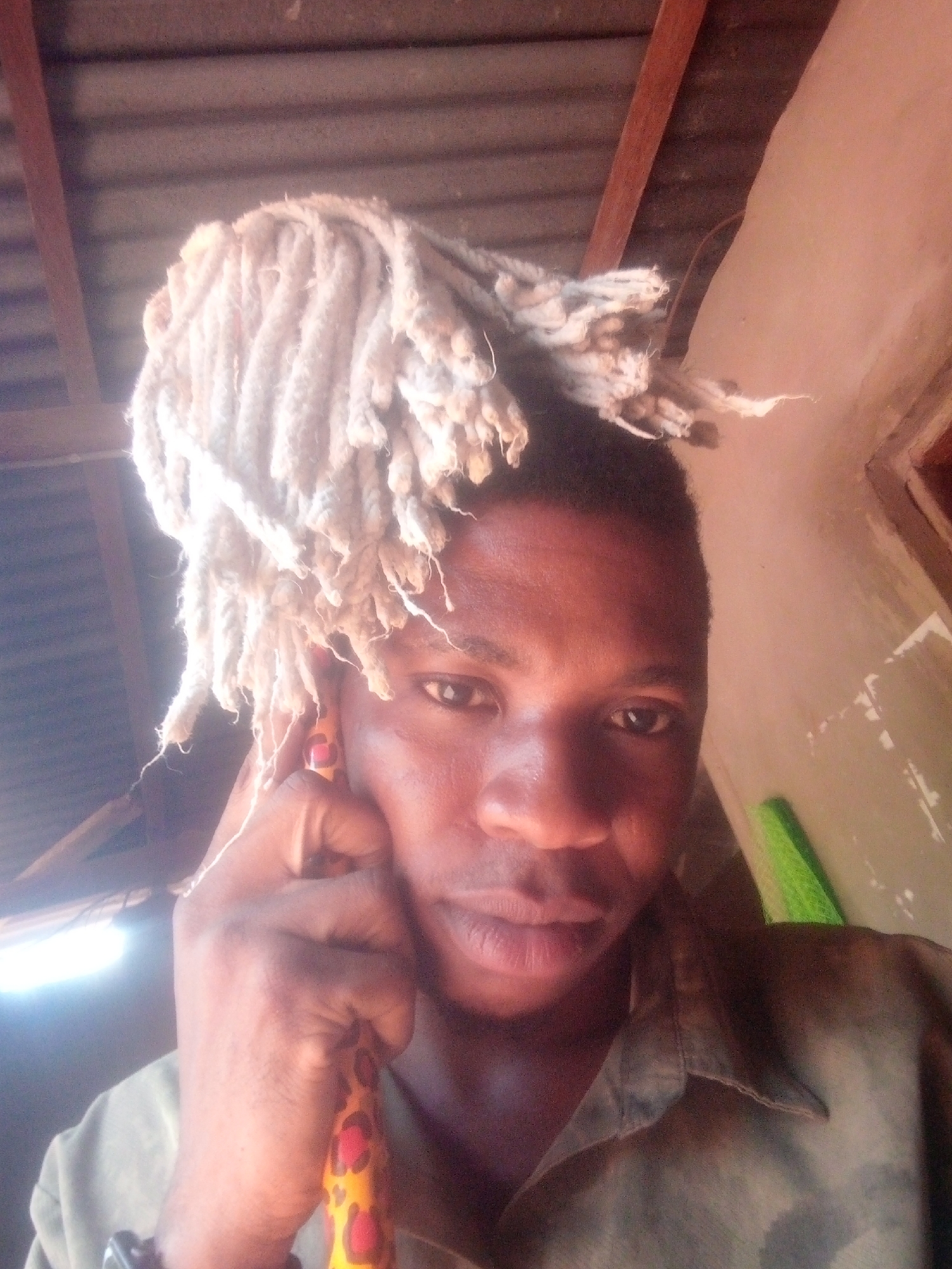 A close-up shot shows a dark-skinned man with short hair dyed a light blonde. His hair is styled in thick, rope-like dreadlocks. He is wearing a collared shirt, possibly olive green in color. His left hand is raised, holding what appears to be a walking stick, possibly made of bamboo, with a patterned fabric wrapping. The location appears to be indoors, with a ceiling constructed of corrugated metal sheets and wooden beams. The exact city and location are unknown.