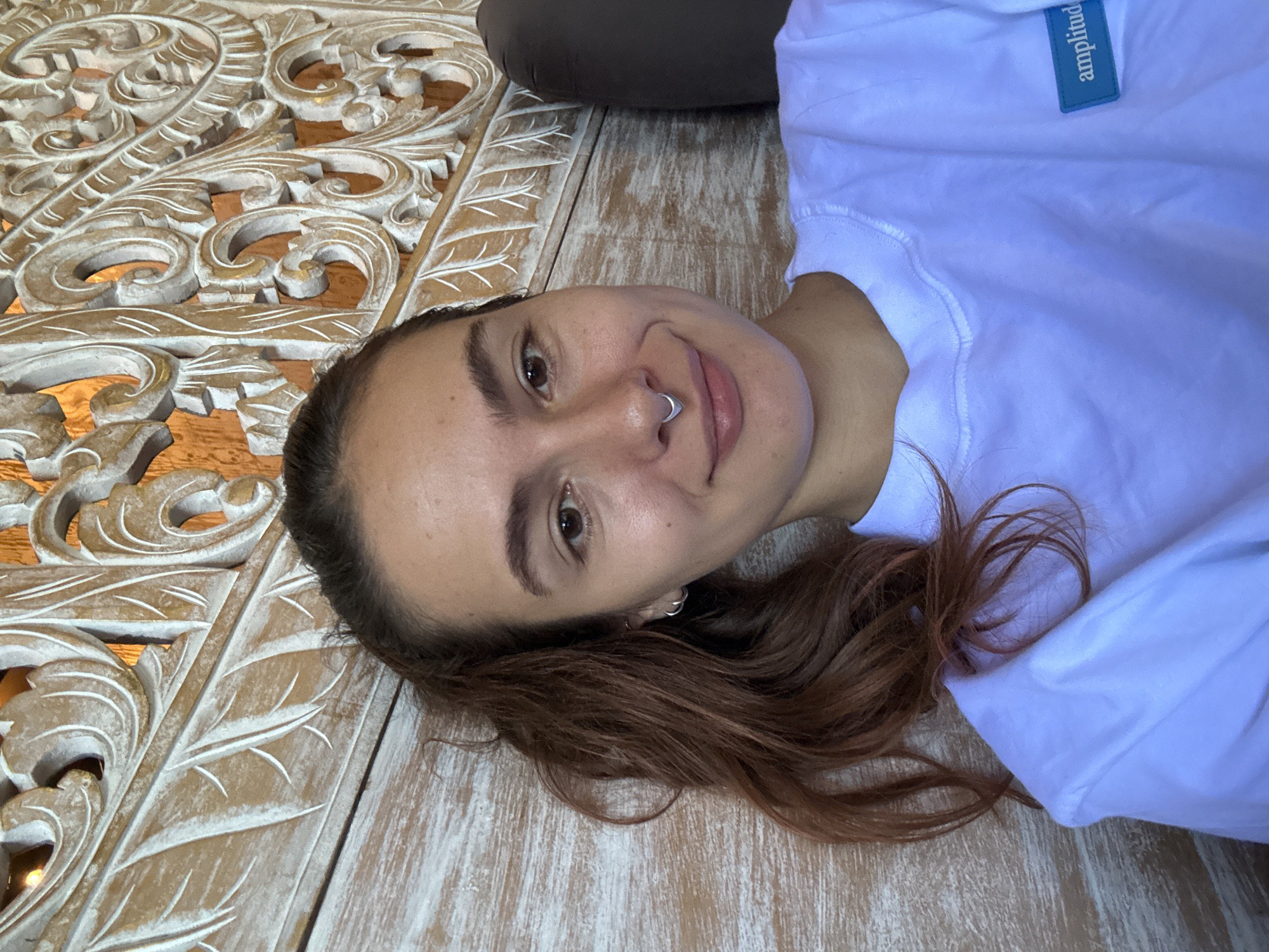A woman lies on a decorative wooden surface, possibly a bed or platform, indoors. The woman is light-skinned, with brown hair, and a nose ring. She is wearing a white shirt with a blue rectangular label bearing the text "amplitude." Part of a brown pillow or cushion is visible at the top edge of the frame. The wooden surface has intricate carved designs. The location is plausibly Canggu, Indonesia, based on the style of carving and general aesthetic.
