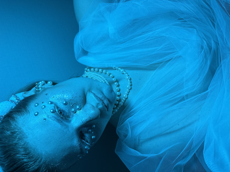 A female subject is depicted in a close-up portrait, illuminated by prominent cyan lighting. Her head is tilted downwards and slightly to the viewer's left, with her gaze directed away from the camera. Her face features elaborate cosmetic adornments: numerous small, pearlescent beads are applied around her eyes and across her upper cheeks, alongside shimmering glitter. Her eyebrows appear lightened. She wears multiple strands of pearl necklaces, including a choker, and several pearl-like earrings or ear cuffs on her visible right ear. Her hair is styled back, partially covered by a light-colored decorative hairpiece. The subject is draped in voluminous, translucent, light-colored fabric, resembling tulle, covering her shoulders and upper torso. The background is a plain, uniformly lit cyan surface. The scene is located in the city of Kazan, Russia.