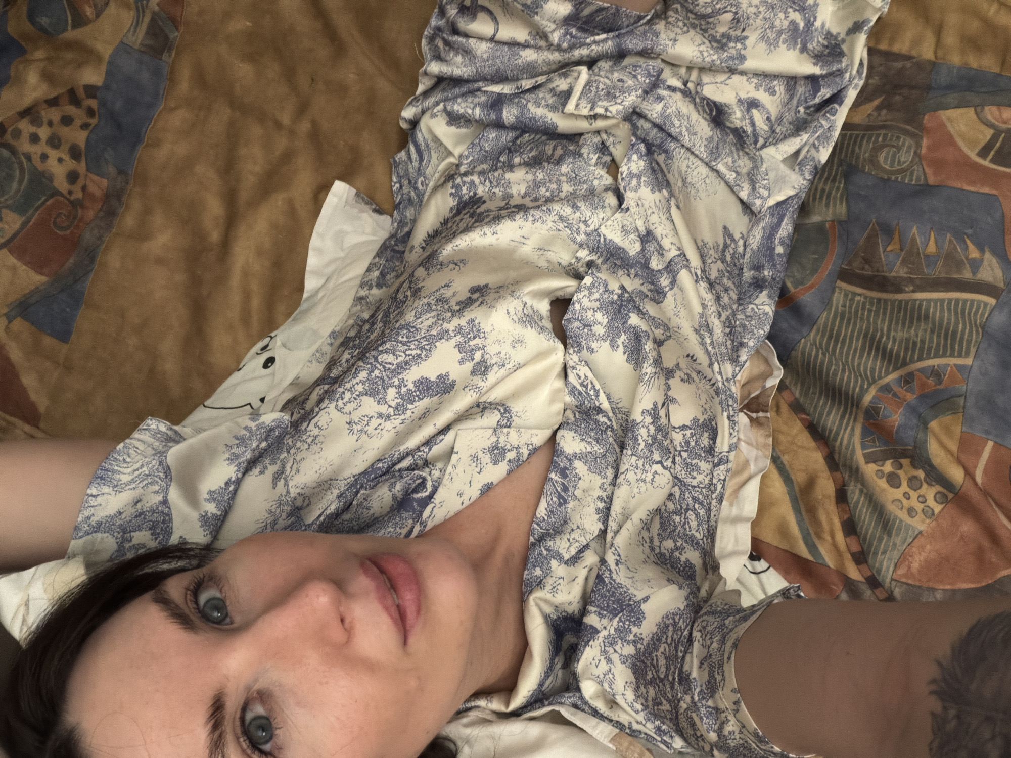 The image is a high-angle, selfie-style photograph of a fair-skinned woman with dark hair and blue eyes. She is lying down on what appears to be a bed or surface covered with multiple textiles. The woman is wearing a white and blue patterned robe or shirt that is partially open at the chest. The background consists of a plain brown fabric on the left and a more colorful fabric with geometric shapes on the right. The photograph appears to be taken indoors. There is no information available to verify the location in the city of Perm, Russia.
