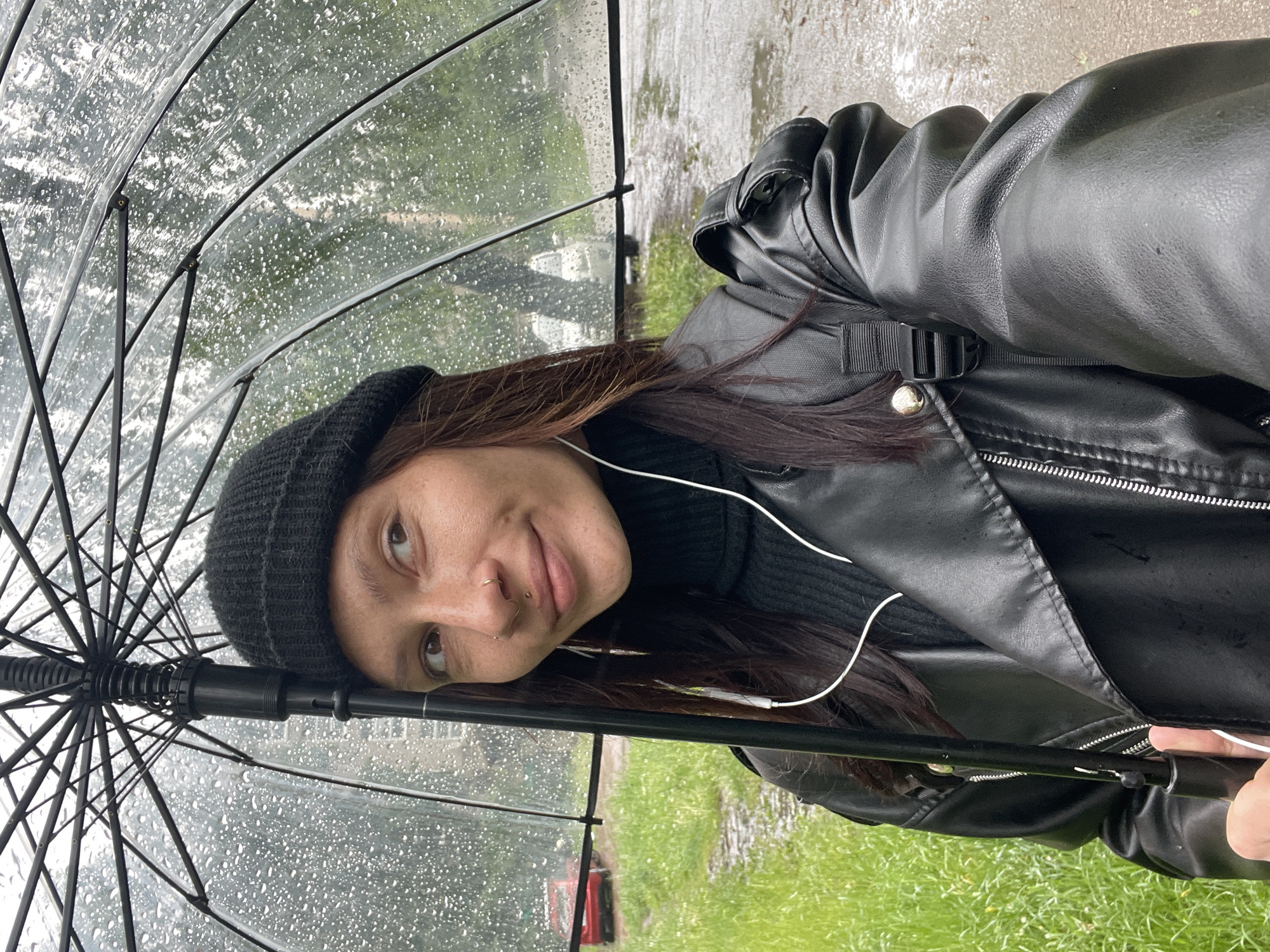 A young woman is standing in the rain in what appears to be an outdoor setting. She is holding a transparent umbrella, with raindrops visible on its surface. The woman is wearing a black beanie, a black turtleneck sweater, and a black leather jacket. She has dark hair and is wearing earphones. A part of her nose ring is visible. The background shows a blurred landscape, suggesting a grassy area and a wet pavement, possibly a park or a street in rainy weather. The image is taken at an angle, giving a different perspective of the scene. There is no other available information. Therefore, the location cannot be determined, however, there is no way to confirm the location is Izhevsk, Russia.
