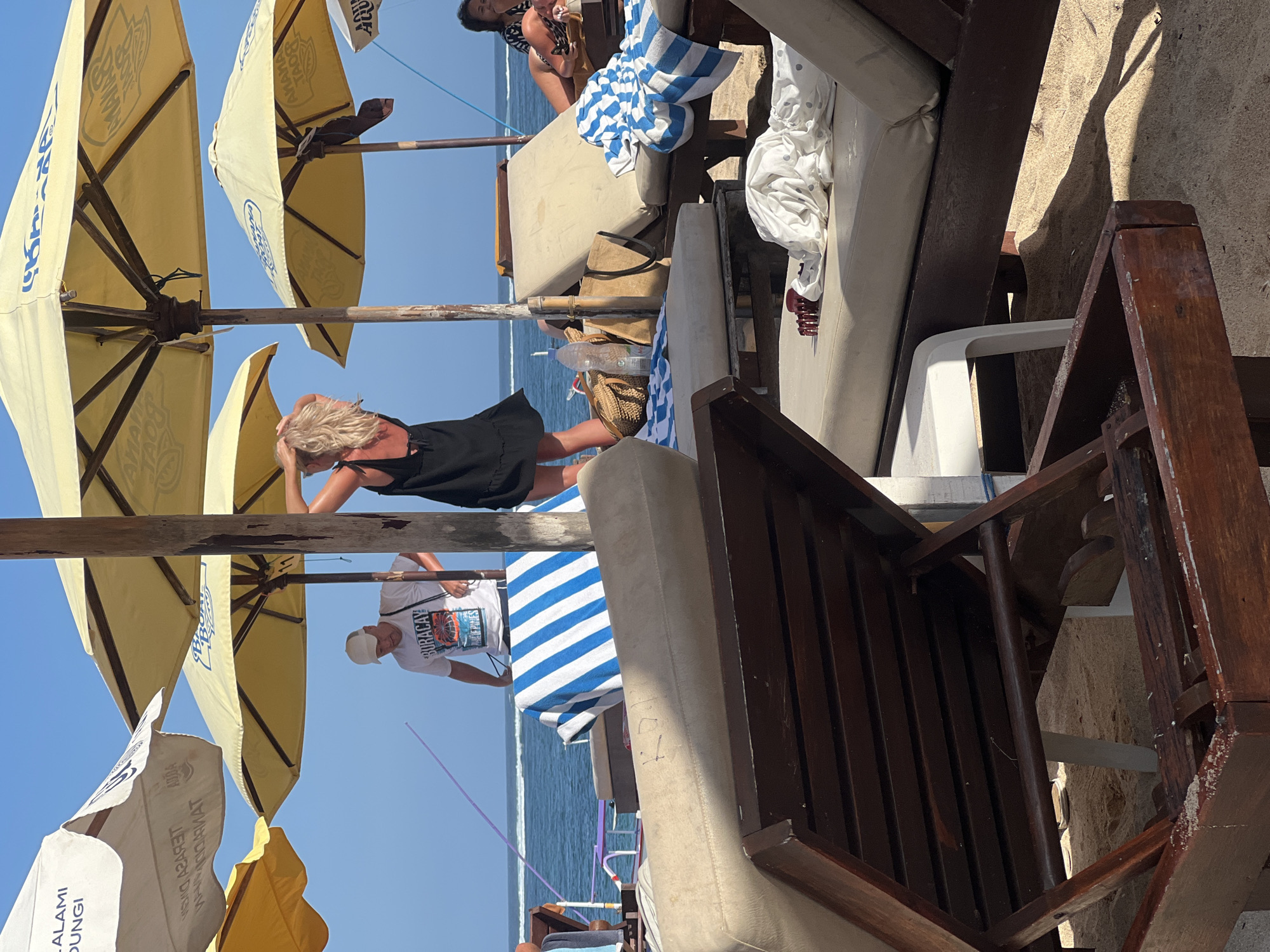 A bright, sunny day at a beach in Kuta Selatan, Indonesia, is depicted. In the foreground, two wooden beach chairs are visible, one empty and partially obscured by the other. The sand is light brown, with some scattered debris on the chairs and sand.
In the mid-ground, several yellow and white beach umbrellas provide shade over a lounge area. Underneath these umbrellas, a person wearing a white t-shirt with the word "BURACAY PHILIPPINES" printed on it is visible, along with a blue and white striped towel laid out on a lounge chair. Another person, a woman with blonde hair, is wearing a black dress and standing with her hands near her head. A woven bag and a water bottle are also present.
In the background, the blue ocean stretches to the horizon under a clear sky. Further back, another lounge chair is occupied by a person wearing a black and white patterned top. The overall scene suggests a relaxed atmosphere at a popular beach destination.