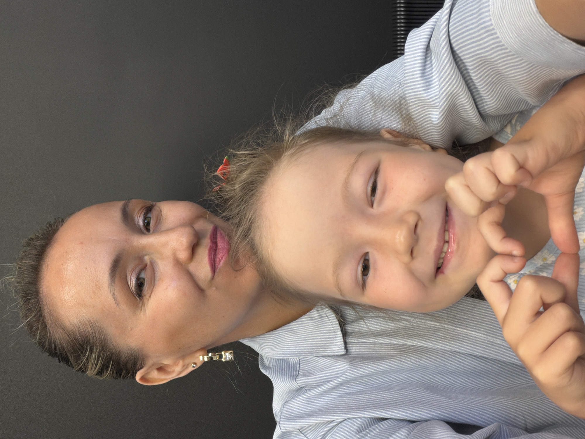 A woman and a child are pictured in a close-up, selfie-style photograph. The woman, positioned behind and to the left of the child, has her hair pulled back and is wearing makeup and a striped shirt. She has her arm around the child. The child, in the foreground, is smiling and making a heart shape with their hands. They are wearing a light-colored shirt with a floral pattern. The background is a dark, solid color. There is no specific information to determine the location in Izhevsk, Russia.