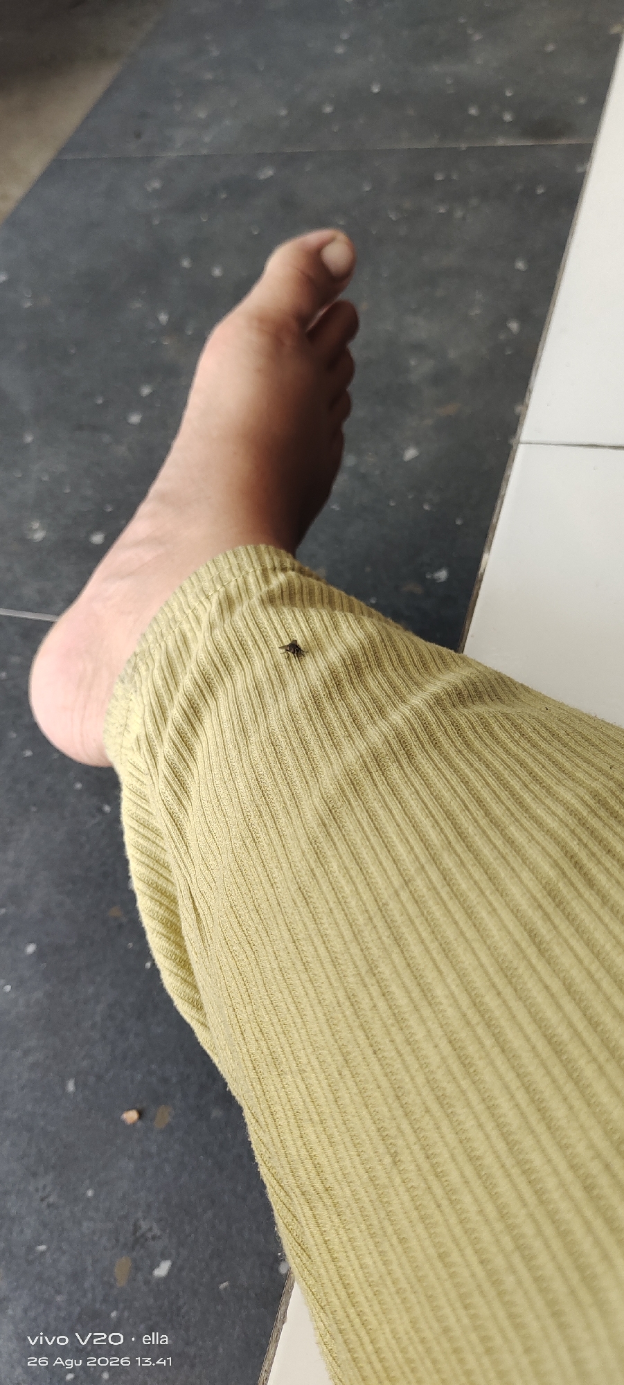 A close-up, overhead shot features a human foot, bare and resting on a dark, speckled floor. The foot is positioned on the left side of the frame, with the toes pointing towards the top. A light yellow, ribbed fabric covers the lower leg, extending from just above the ankle to the edge of the frame. A small, dark insect, likely a fly, is visible on the fabric, approximately in the center of the image. The background is a mix of dark flooring and a sliver of white material on the right. The timestamp "vivo V20 · ella 26 Agu 2026 13.41" is present in the bottom left corner, indicating the photo was taken at 13:41 on August 26, 2026. The location is specified as Simo, Indonesia.