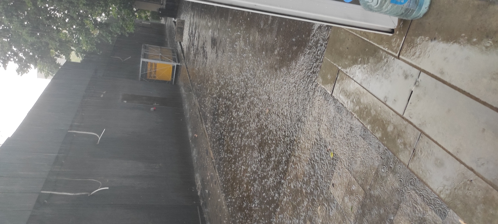 The image depicts a street scene in Kamianske, Ukraine, during a rain shower. The ground is wet, with visible ripples from raindrops on the pavement. A dark, textured wall runs along the left side of the frame, with some white conduits attached to it. To the right, a tree with green foliage is visible, also being affected by the rain. In the middle ground, there is a caged structure containing a bright yellow object. Further to the right, a white door with a red handle is set into the wall. A portion of a white doorframe or window frame is visible on the far right edge of the image, along with a large plastic bottle of water, also wet from the rain. The overall impression is of an urban environment experiencing inclement weather.