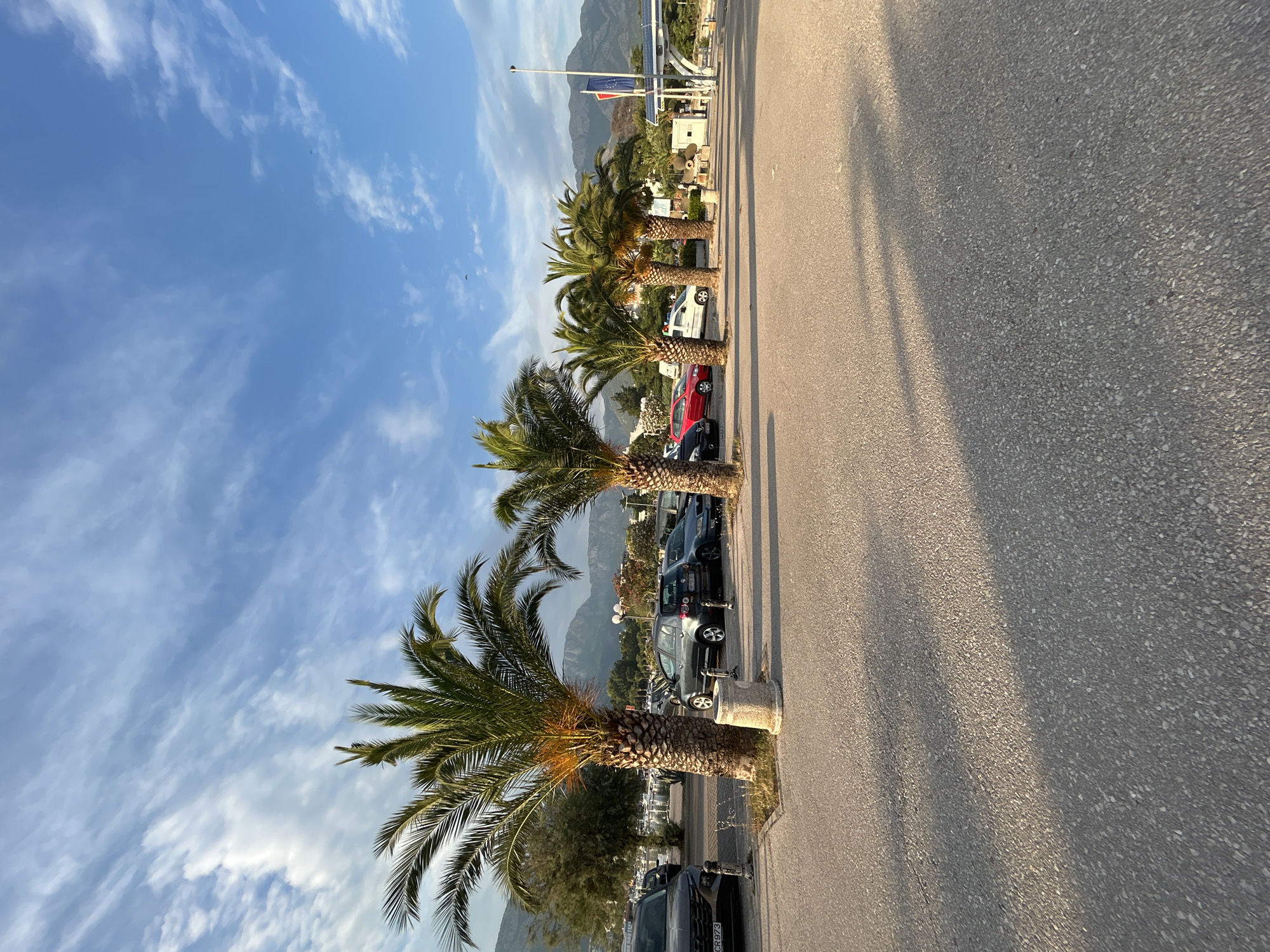 The image shows a roadside scene with palm trees lining a paved area. In the background, there are mountains under a bright blue sky with wispy white clouds. Several cars are parked along the roadside. Some of the cars are dark-colored, and one is red. One of the cars is a dark gray hatchback. There are also some buildings and structures visible in the distance, including what appear to be solar panels and flags. The sun is casting long shadows on the pavement, indicating it is likely late afternoon or early evening. The overall impression is a warm, sunny day in a coastal or Mediterranean-like location. The presence of palm trees, mountains, and the sea (implied by the parked boats in the distance) suggests a scenic, potentially tourist-oriented setting. No people are visible in the image. Some text is visible on a license plate of a dark car in the foreground, appearing to be "CR973".