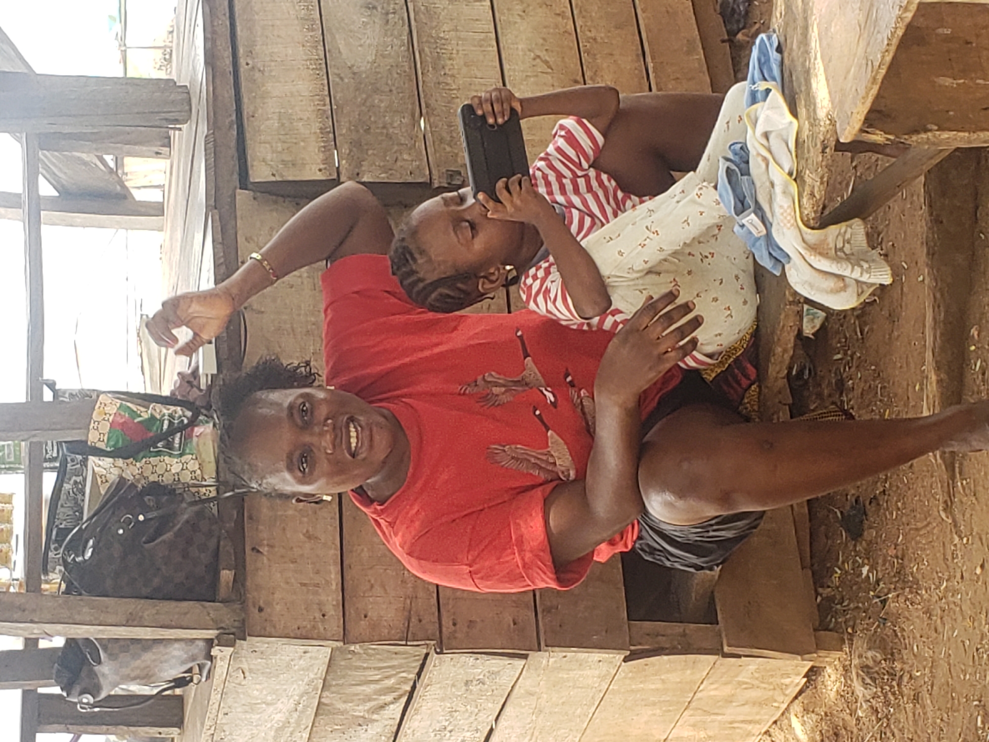 A woman and a child are visible on a wooden structure with an open dirt space beneath. The woman is wearing a red t-shirt and is seated. A child in a striped shirt and patterned pants sits with the woman, holding a tablet. A purse hangs on the structure to the left. The location is presumed to be Adankolo, Nigeria, based on given data.