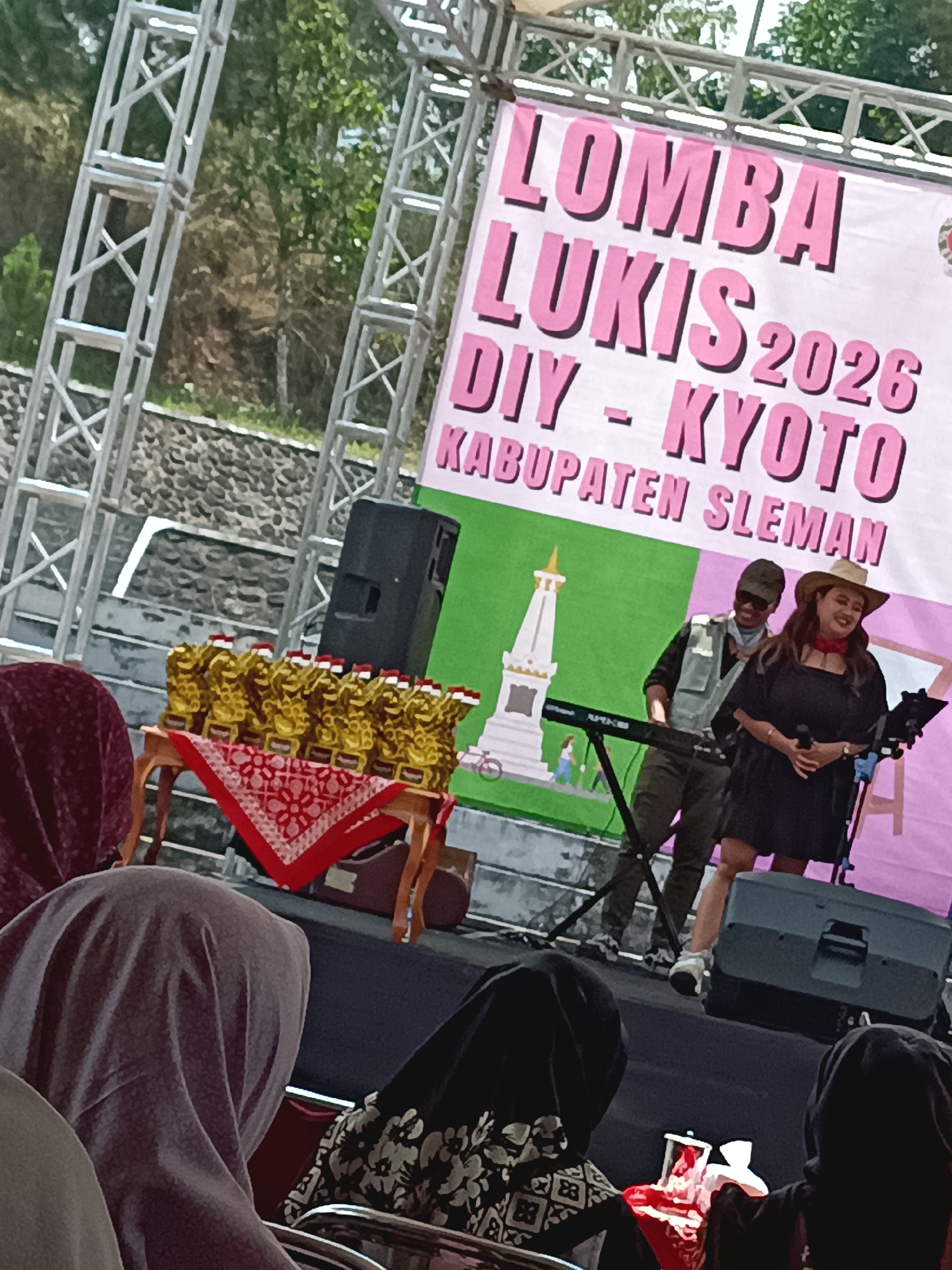 The image shows an outdoor event stage in Hargobinangun, Indonesia. A large pink banner with Indonesian text, likely advertising the event, is visible in the background. The text reads "LOMBA LUKIS DIY - KYOTO 2026 KABUPATEN SLEMAN". Two performers are on stage: a woman wearing a black dress and a cowboy hat, holding a microphone and singing, and a man in a vest and sunglasses operating a keyboard. In the foreground, an audience is seated, many of whom are wearing headscarves. A table with several golden trophies is also displayed on stage. The overall scene suggests a cultural event or competition.