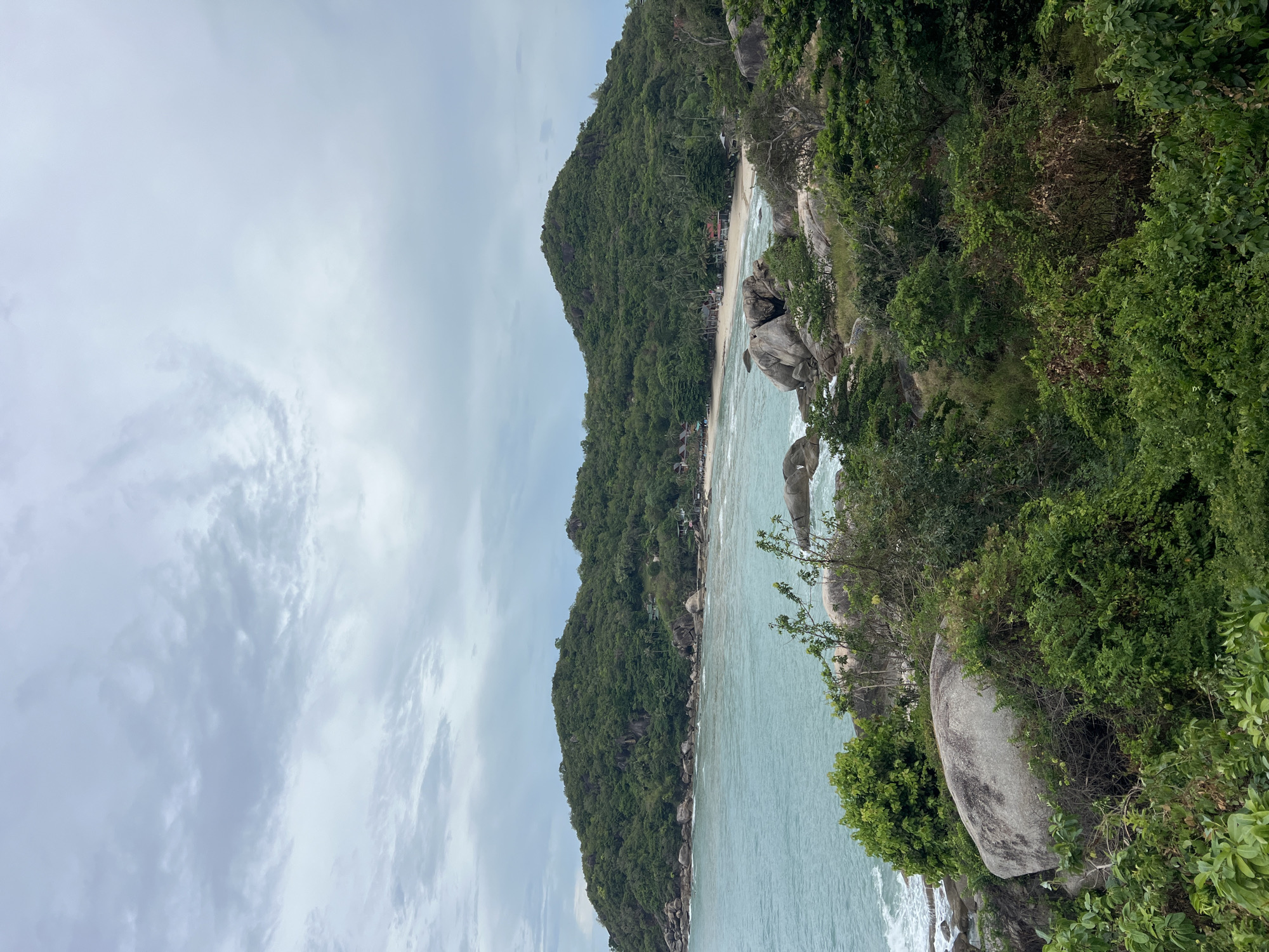 The image shows a coastal scene in Baan Chaweng Noi, Thailand. A densely vegetated, green hillside descends towards a sandy beach, which borders a body of light-blue water. Large rocks are scattered along the shoreline. The sky is overcast with grey clouds. The viewpoint is elevated.