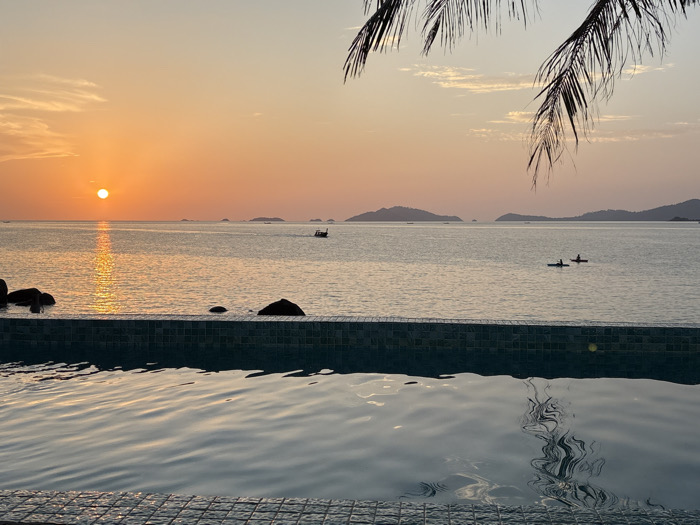 An outdoor scene captured at sunset in Ban Ko Li Pe, Thailand, features an infinity pool foregrounding a calm sea. The pool's edge, constructed with small dark mosaic tiles, occupies the lower third of the frame, its water reflecting the sky and exhibiting gentle ripples. The sun, a prominent orange disc, is positioned just above the horizon on the left, casting a shimmering path of light across the water. Several large, dark rocks are partially submerged in the sea near the pool's left side. In the midground, two individuals in kayaks or small boats are visible on the water towards the right, while a traditional longtail boat moves horizontally further to the right. Distant islands or landmasses are silhouetted against the horizon. The sky displays a gradient of warm orange, pink, and light blue hues, with sparse, wispy clouds. Dark palm fronds intrude into the upper right corner of the frame.