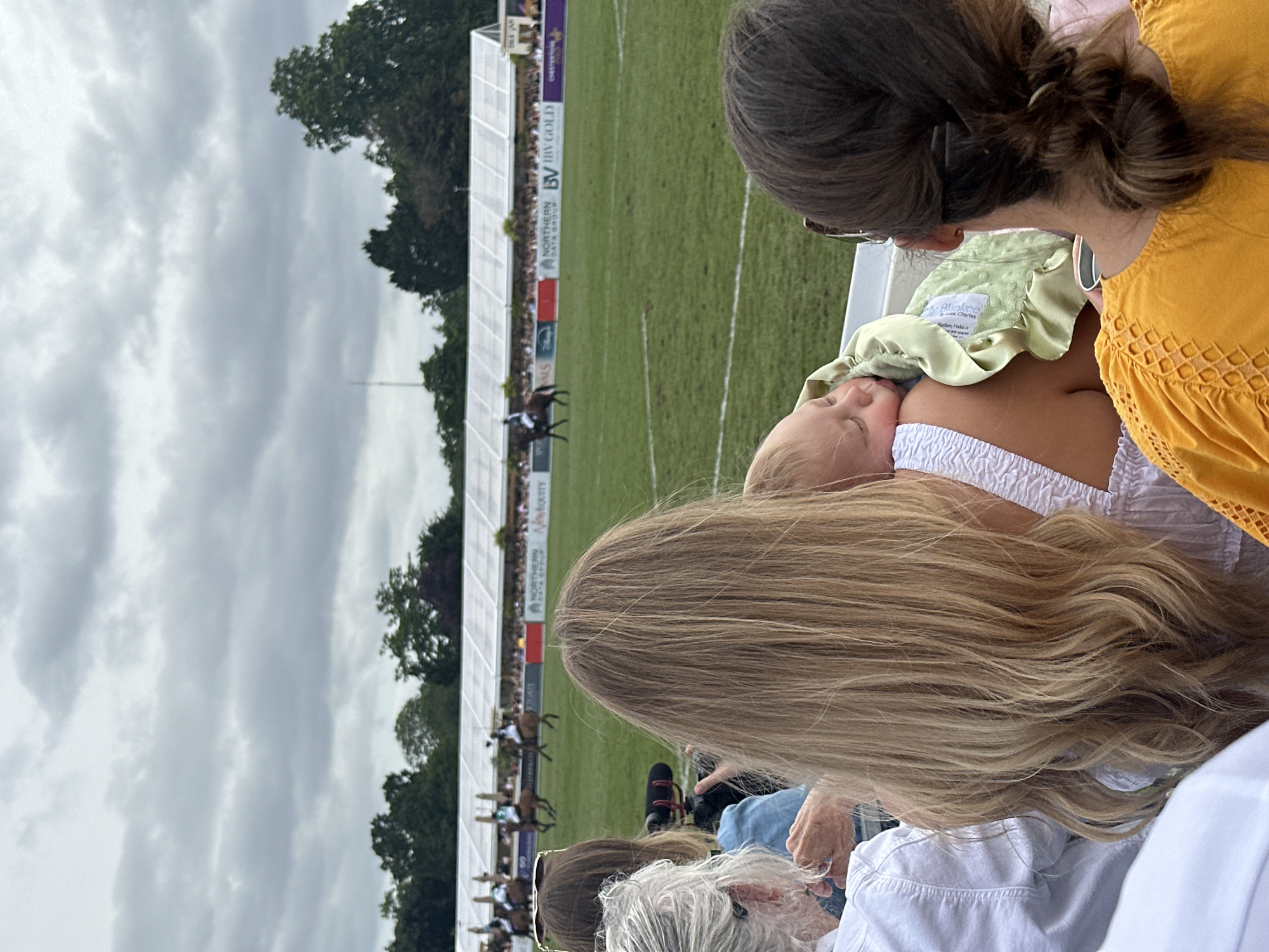 Four individuals are observed from behind in the foreground, seated or standing, facing a grassy field. On the far left, an individual with short, wavy grey hair, wearing a white collared shirt, is partially visible. Centrally, a female with long, wavy blonde hair, wearing a white ruched-strap top, holds an infant on her left shoulder. The infant, wearing a light-colored garment, is swaddled in a light green textured blanket and appears to be sleeping, with its head resting on the woman's shoulder, facing away from the camera. To the right, a female with dark brown hair styled in a low ponytail, secured by hair clips, wears a mustard-yellow top with textured detailing on the shoulder; sunglasses rest on top of her head. Partially obscured behind the grey-haired individual, another person holds a black camera with an attached external microphone, pointed towards the field.
The midground features a large, green sports field marked with white lines, where several horses with riders in light-colored uniforms are observed in motion. A long, white marquee structure spans the far side of the field, backed by a crowd of spectators. Below the marquee, a series of sponsor banners are visible, displaying logos and text including "Sport," "Disney," "NORTHERN DATA GROUP," "BV BV GOLD," and partially visible "New Equity" and "Chester Polo."
The background consists of a dense line of green trees beneath an overcast sky with grey clouds. A thin, vertical structure, possibly an antenna or mast, is faintly visible above the trees on the left side of the frame. Based on the "Chester Polo" banner visible in the midground, the event is likely taking place in Chester, United Kingdom. The image does not contain specific visual identifiers for the city of London, United Kingdom.