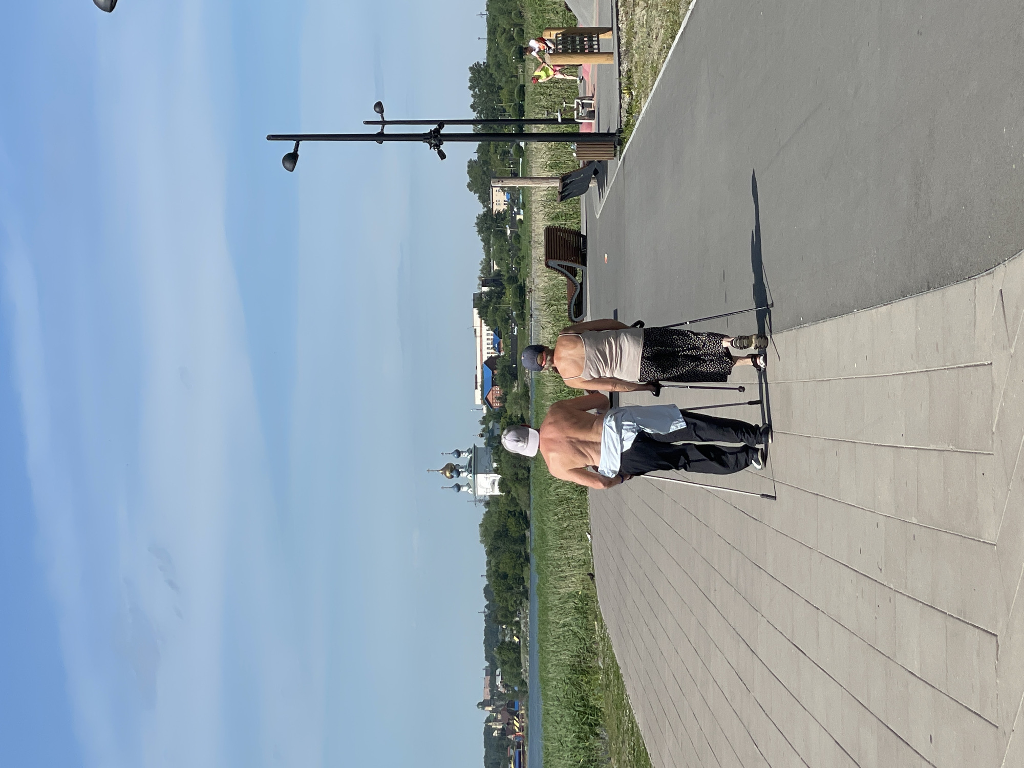 Two individuals, a man and a woman, are depicted from behind as they engage in Nordic walking along a paved pathway in a park. The man, bare-chested and wearing black pants and a white cap, carries a light blue garment tied around his waist. The woman, wearing a beige tank top and a patterned skirt, also uses Nordic walking poles. They are walking away from the camera, towards a body of water visible in the mid-ground. Beyond the water, a town or city skyline is visible, featuring several buildings, including a prominent church with blue domes. The environment appears to be a sunny day with a clear blue sky and some scattered clouds. The pathway is lined with vegetation and includes park amenities such as benches and lampposts. In the background, other individuals can be seen using outdoor exercise equipment.