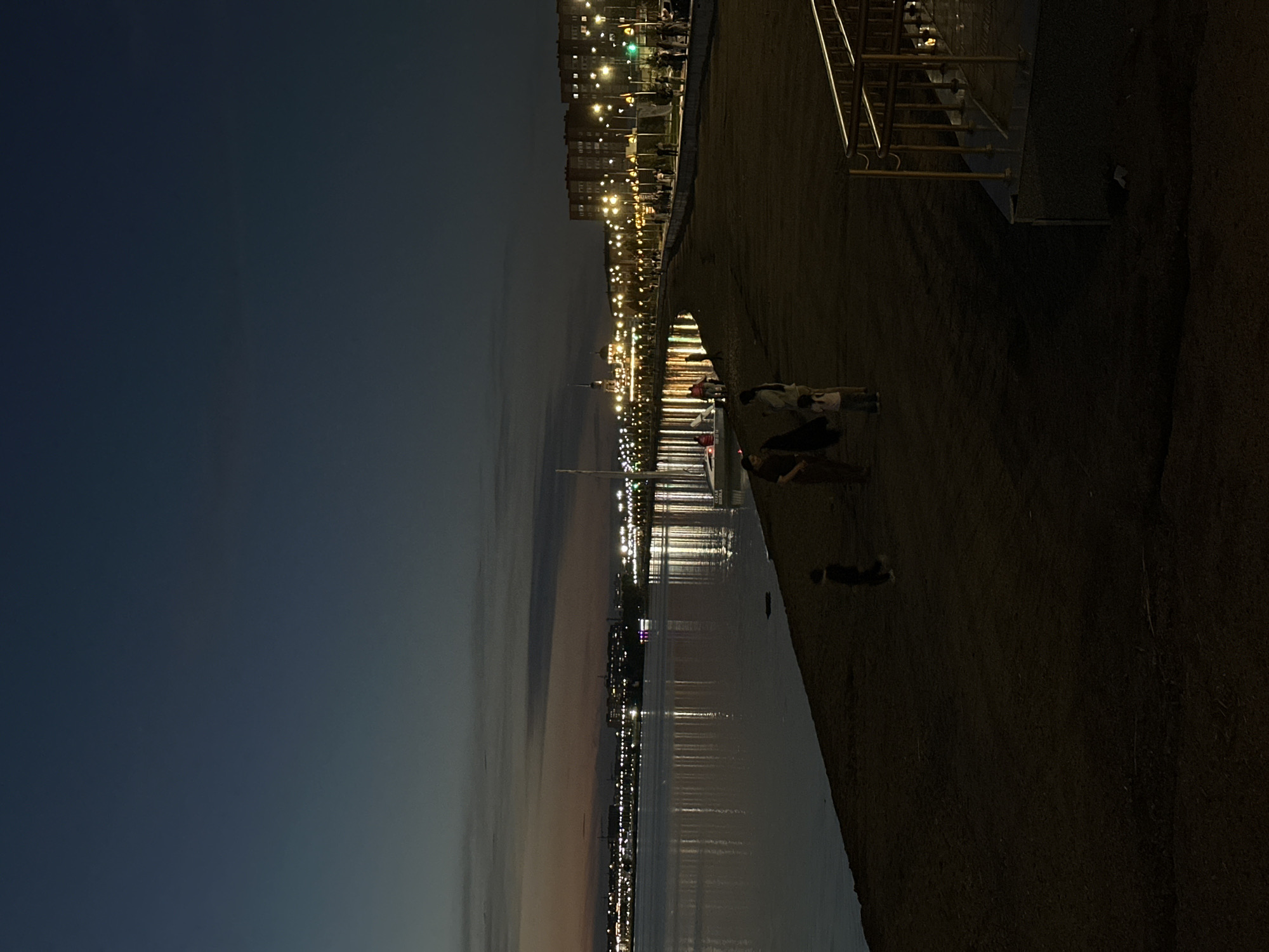 The image depicts a twilight scene at a waterfront, likely in Kokshetau, Kazakhstan. The sky is a deep gradient of blues and grays, indicating either the end of the day or the beginning of the night. A body of water, possibly a lake or river, reflects the numerous lights of a city or town that stretches along the opposite bank. The lights are primarily yellow and white, creating a shimmering effect on the water's surface.
Along the near bank, there is a paved pathway or promenade, bordered by a dark, textured ground that appears to be sand or dirt. A metal railing, likely part of a walkway or stairs, is visible on the right side of the frame.
Several people are present in the scene, though their features are indistinct due to the low light. On the promenade, a few individuals are walking or standing. In the foreground, closer to the water's edge, two people are seated or lying down on the ground. Further down the promenade, there are more figures, suggesting a gathering or evening stroll. The presence of people and the illuminated city suggest this is a public space where people enjoy the evening.
The overall mood is peaceful and serene, with the reflections of the lights creating a visually appealing atmosphere. The time of day is clearly after sunset. There is no visible text in the image.