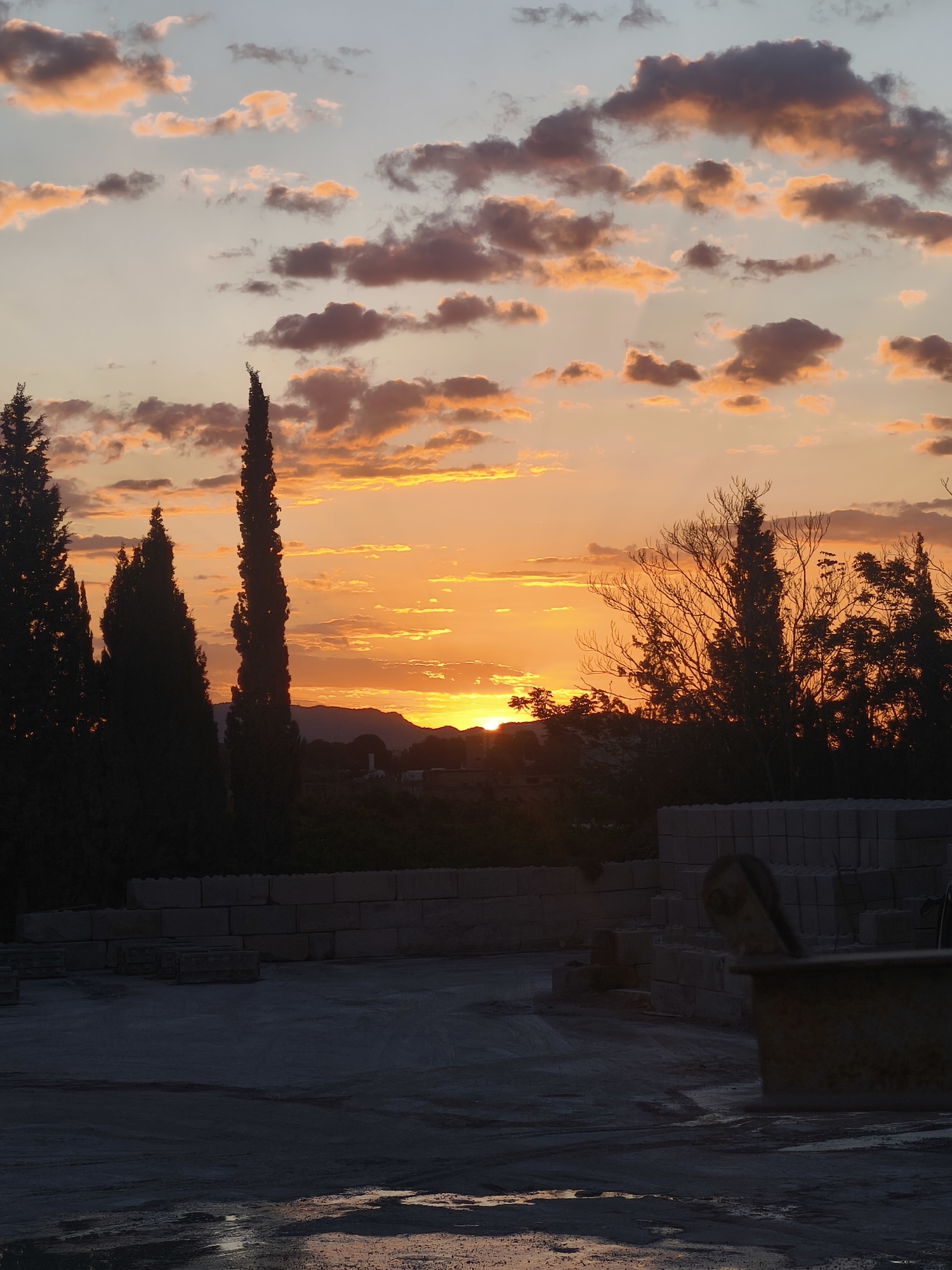 The image captures a sunset scene, likely in an industrial area, with a view towards a distant mountain range. The sky is filled with clouds illuminated in shades of orange and yellow, indicative of a setting sun. In the foreground, there are construction materials, including stacked blocks, and what appears to be a piece of industrial equipment. Silhouetted trees are visible in the midground, partially obscuring the view of the mountains and the setting sun. There is an area of standing water in the foreground, reflecting some of the sky's colors. Given the prompt, the location is likely near Bétera, Spain. There are no people visible in the image. The overall atmosphere suggests twilight or early evening.