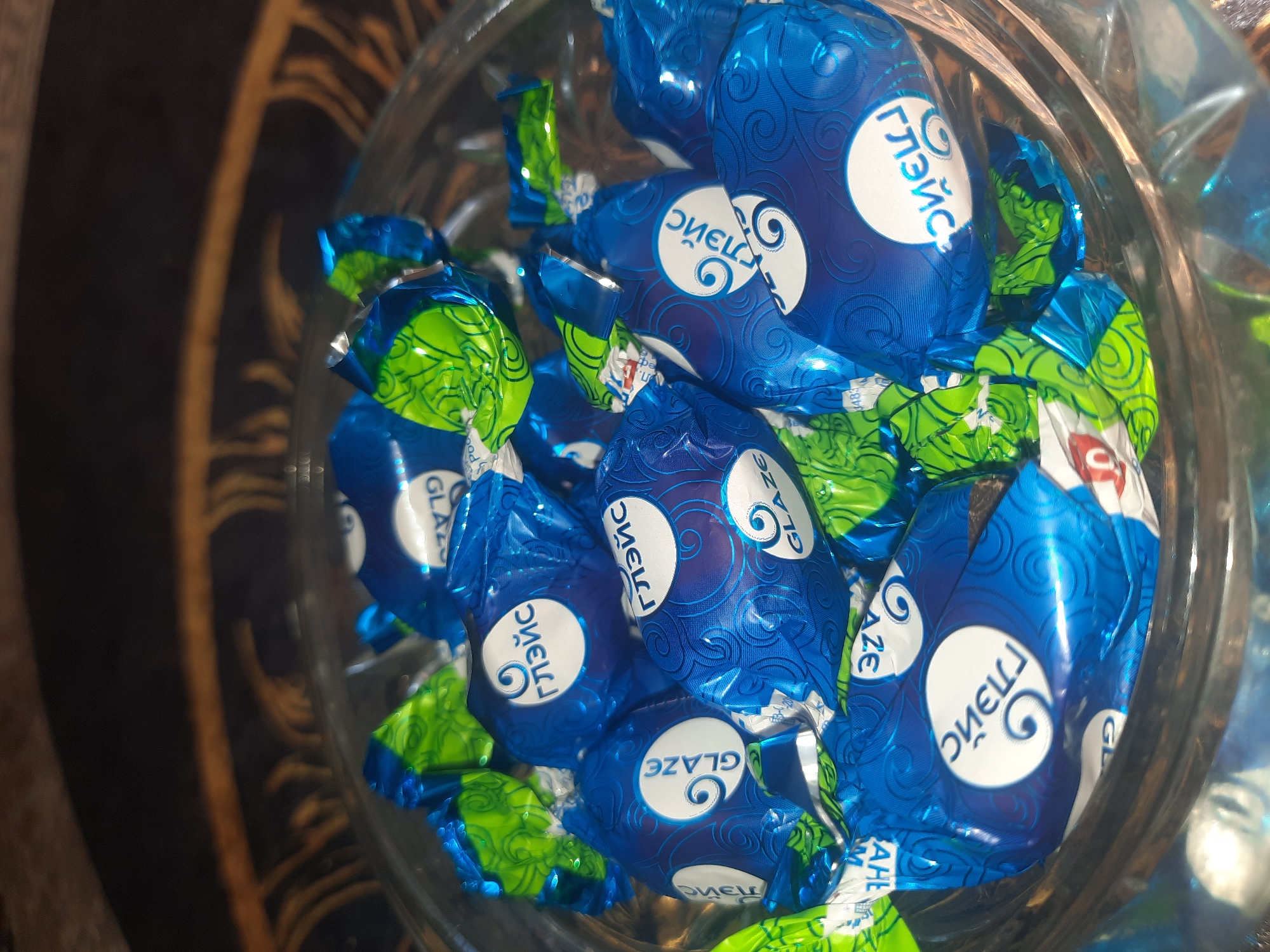 The image shows a close-up, overhead view of a clear glass bowl filled with individually wrapped candies. The candies are predominantly blue and green, with some in a metallic silver wrapper. The blue wrappers feature a white circular logo with a swirling design and text that appears to be in Cyrillic, possibly "Глейс" (Gleys). Some of the blue wrappers also have the word "GLAZE" printed in English. The green wrappers have a swirling pattern. The candies are densely packed in the bowl, with some wrappers creased and others more intact. The background is dark and out of focus, with hints of a patterned surface and possibly some golden trim. The lighting is dim, casting reflections on the candy wrappers. There are no people visible in the image. The scene is indoors, likely in a home or shop, and depicts a collection of confectionery. The location in the city of Шклоў, Belarus is not discernible from the visual information presented in the image.